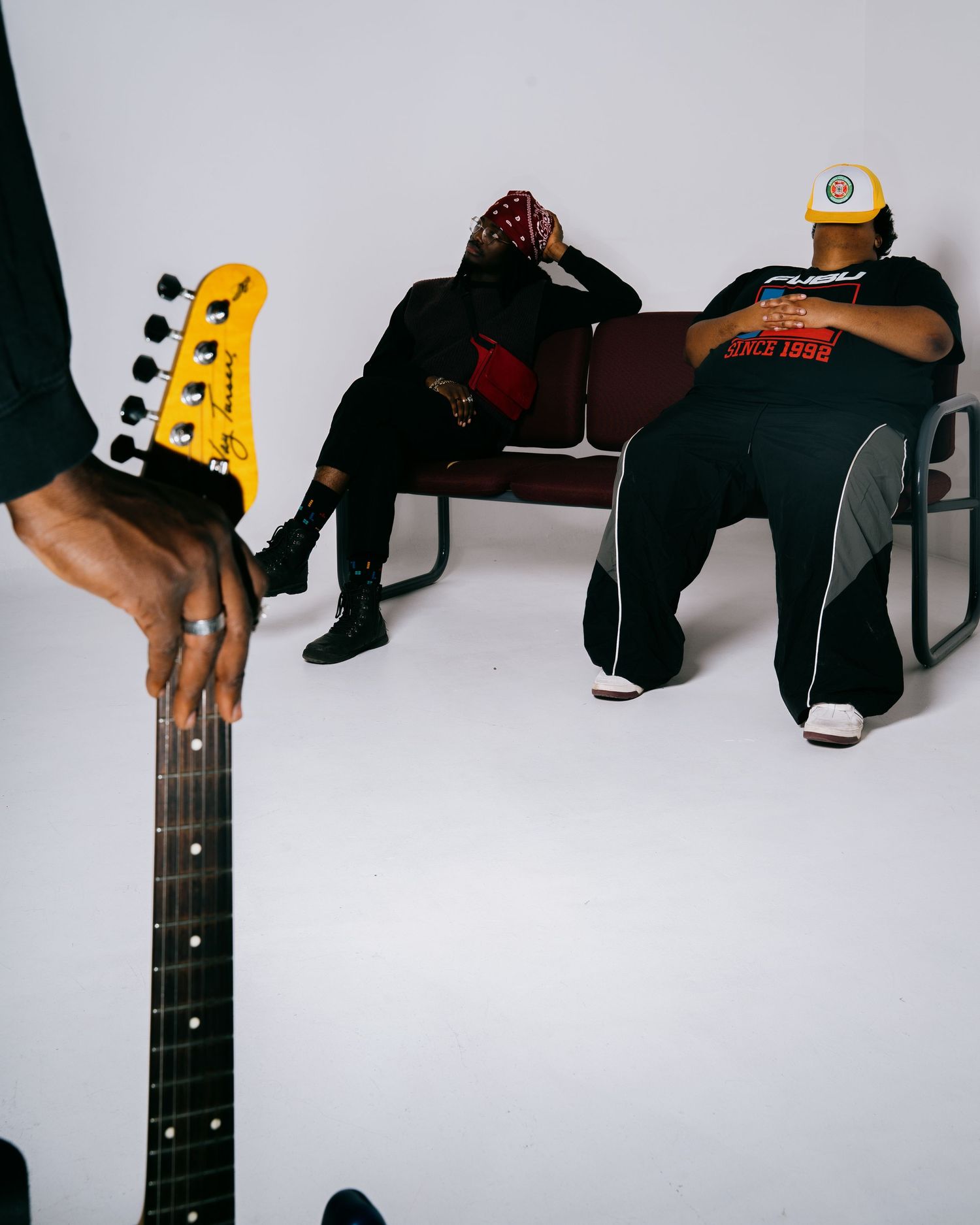 Yellow electric guitar neck in foreground with two musicians resting on bench behind.