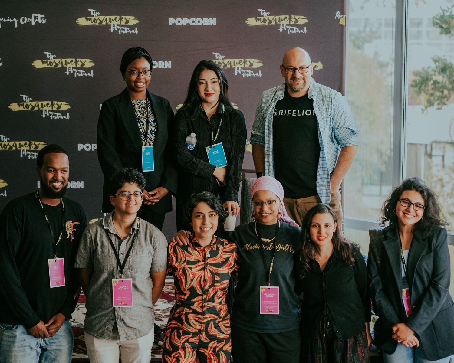 A diverse group of nine people pose together in front of a festival backdrop with lanyards and badges.