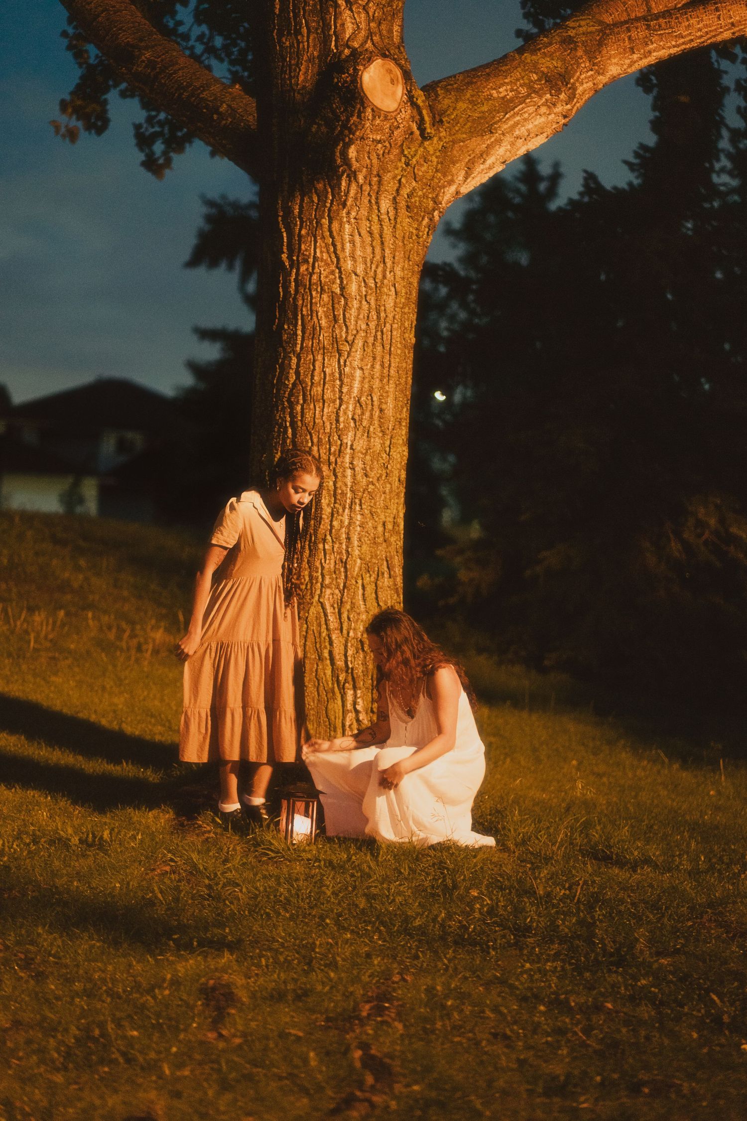Two women sit under a large tree at night, one in white holding a glowing lantern on green grass.