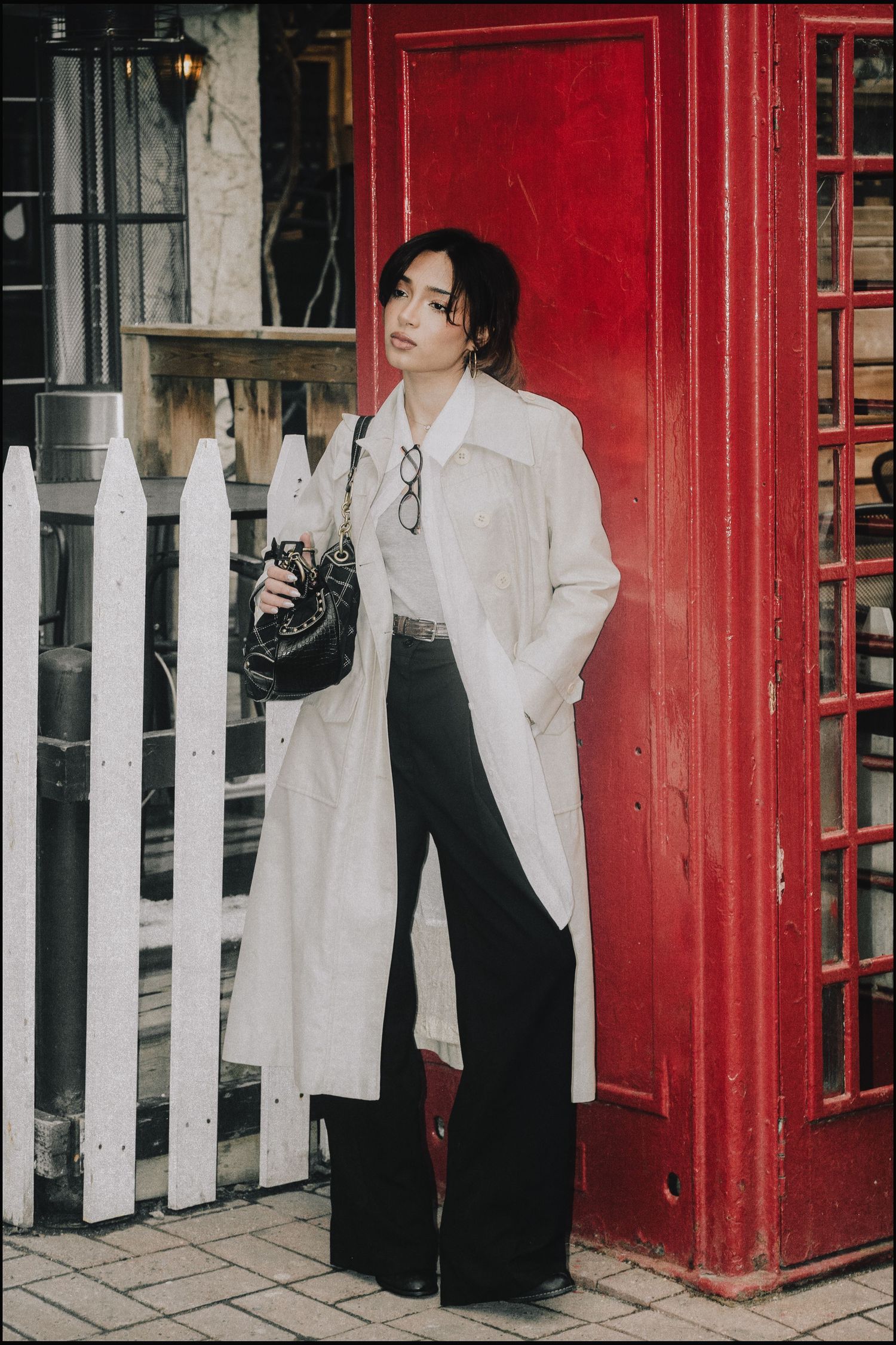 Woman in white trench coat and black wide-leg pants posing by red phone booth.