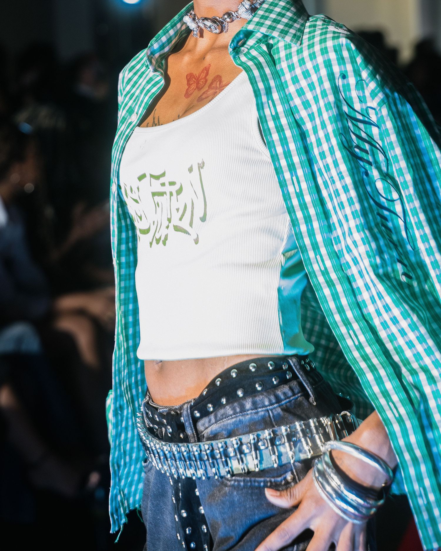 Close-up of model in teal plaid shirt over white graphic crop top with studded denim waistband.