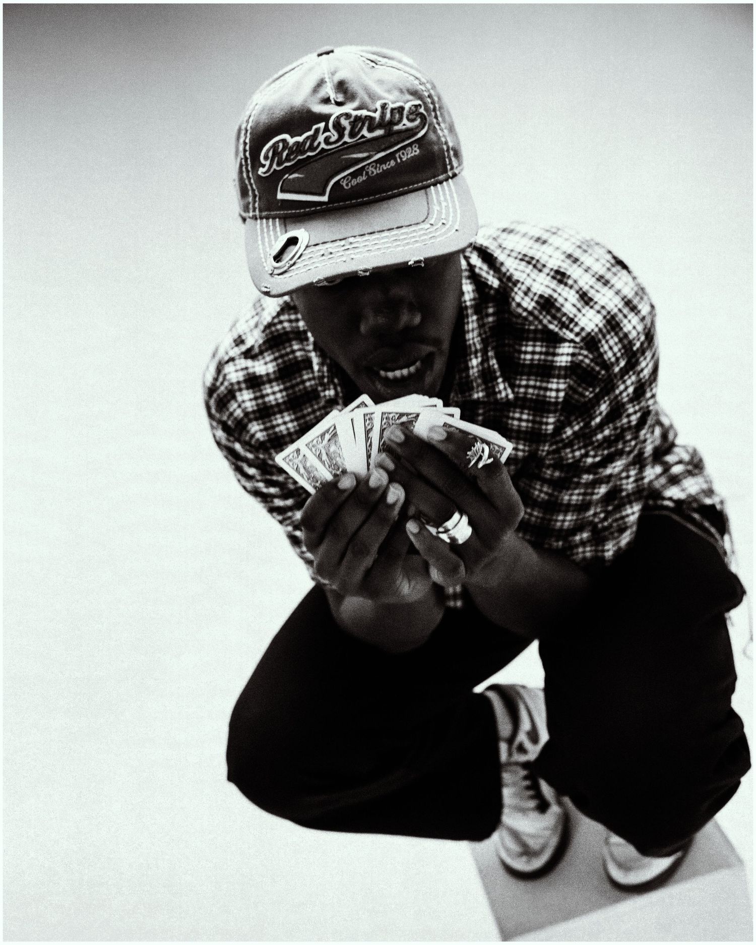 Black and white photo of a man crouching and holding a fan of cash bills wearing a plaid shirt and snapback cap.
