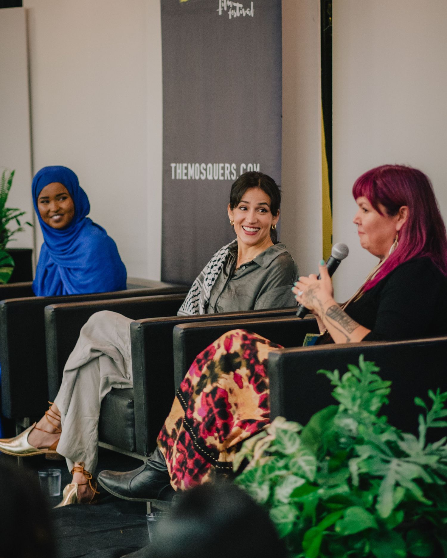 Three women participate in a panel discussion, one holding a microphone, seated in front of a festival banner.