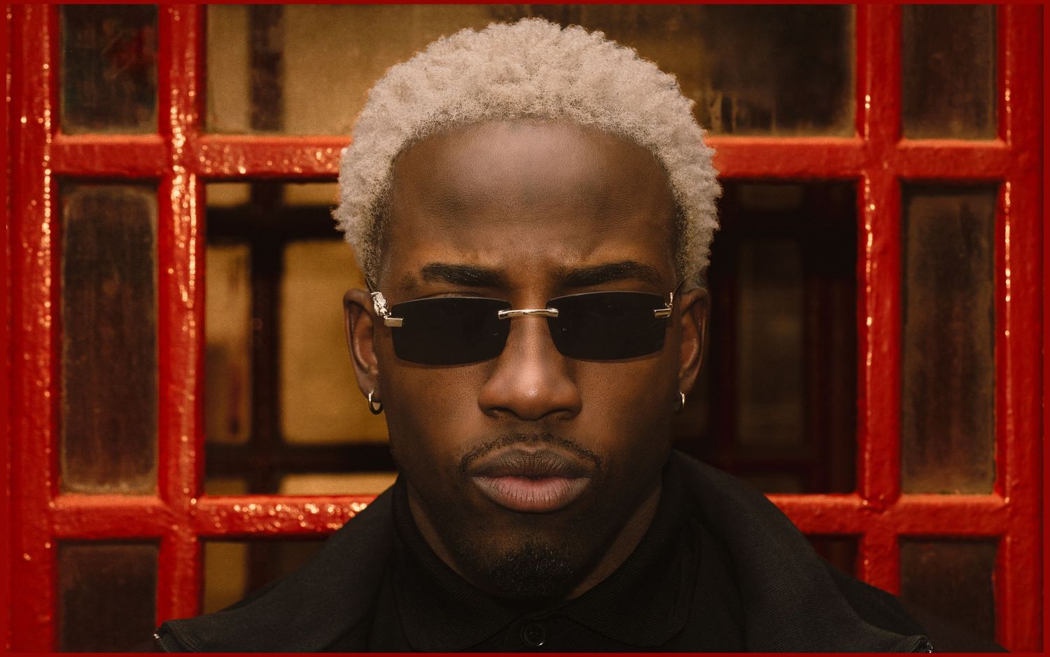 Man with platinum blonde hair and dark sunglasses poses in front of red phone booth.