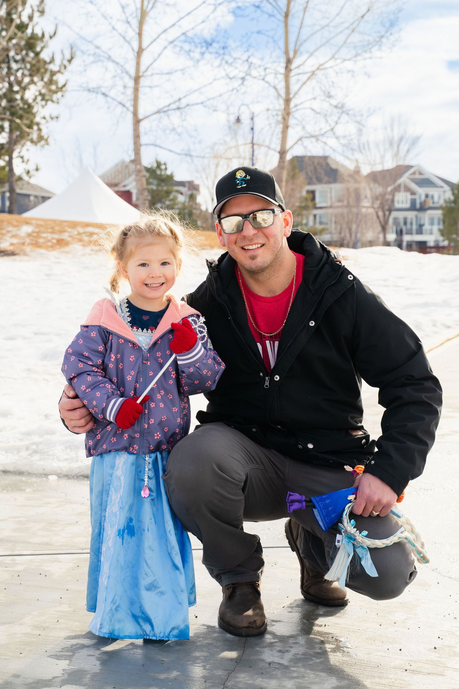 Young girl in Elsa costume posing with smiling man crouching beside her on frozen outdoor ice rink.