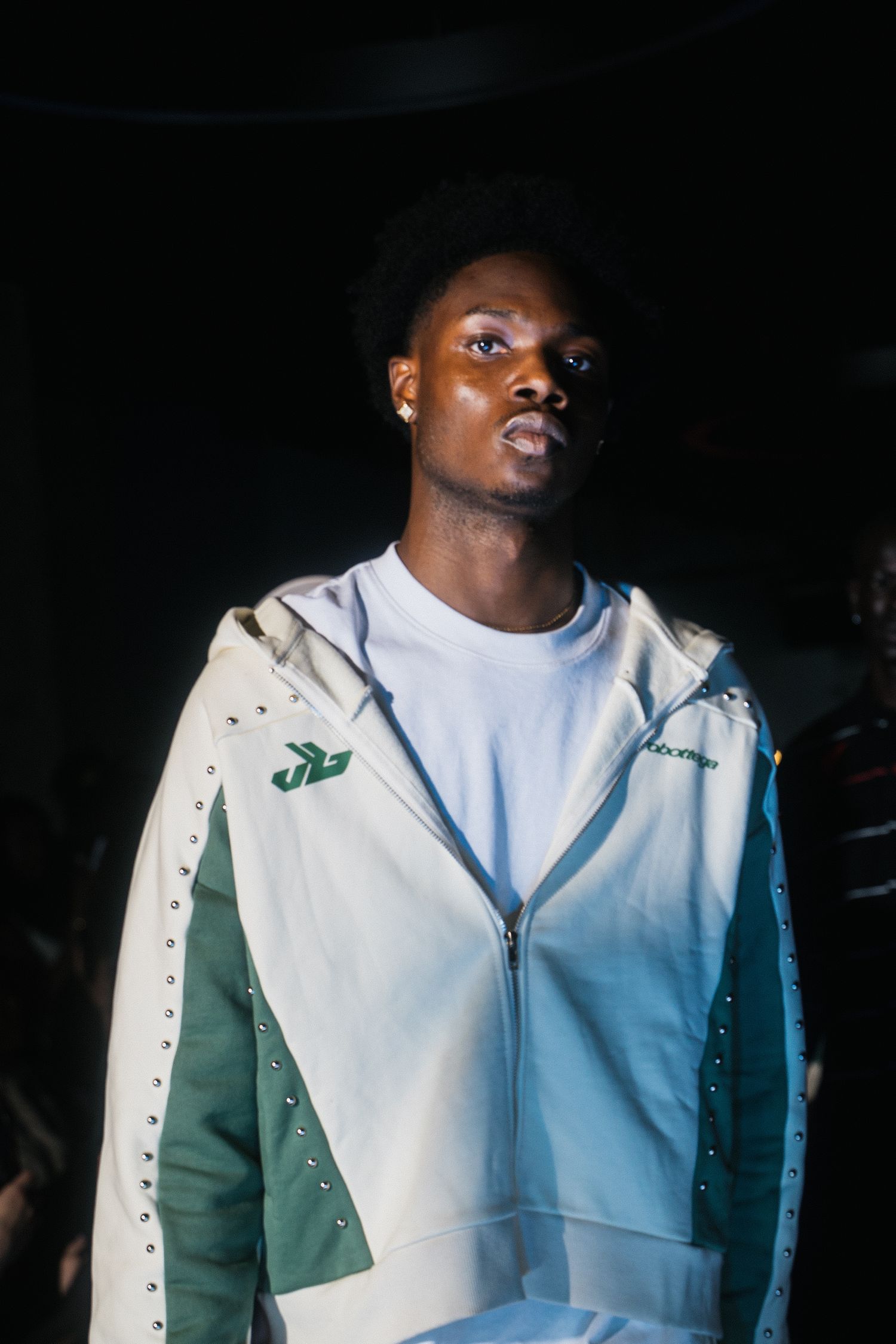 Young male model poses in white and green studded zip-up jacket at an indoor fashion event.
