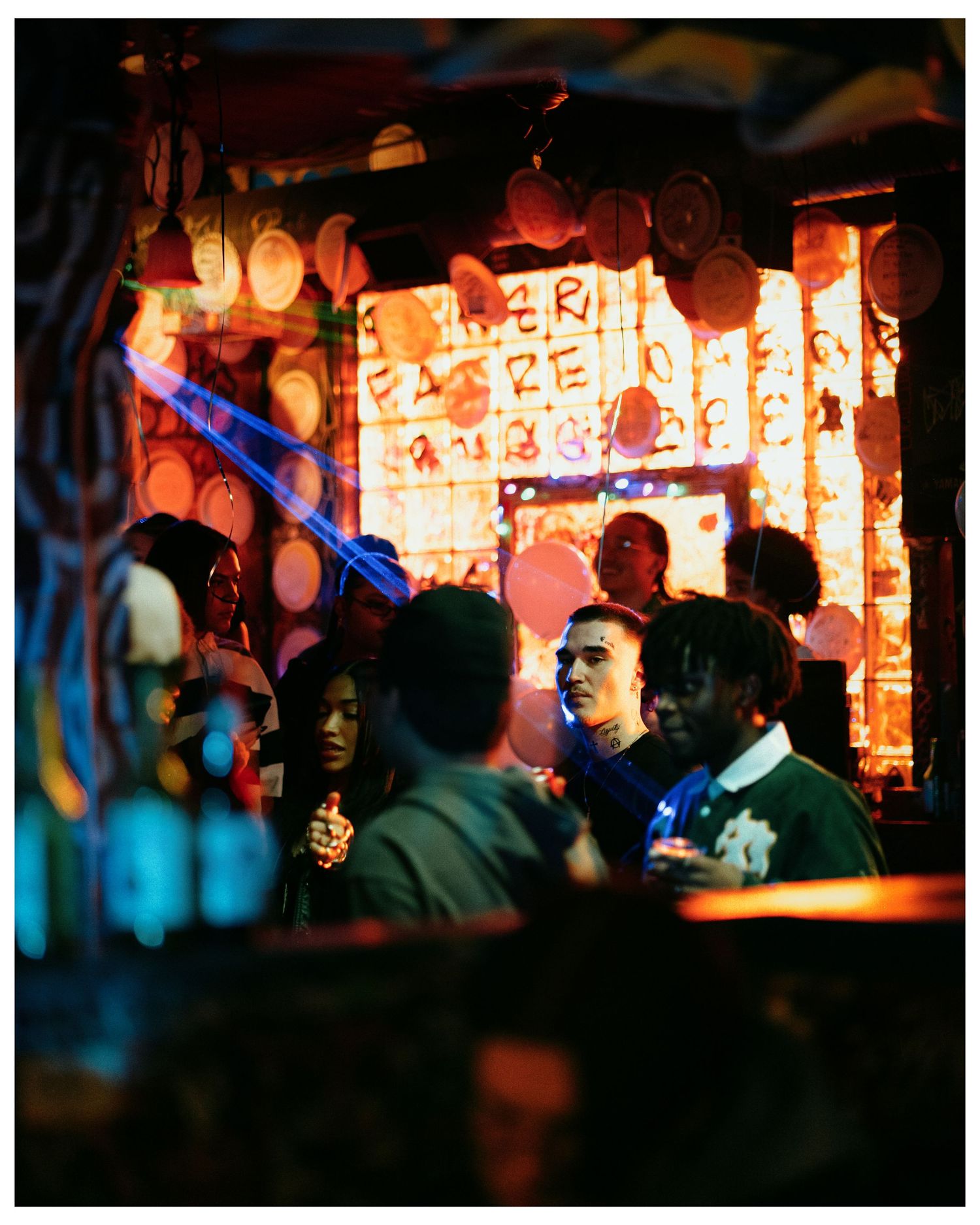 A group of people socialize in a vibrant, colorfully lit bar with glowing orange decor.