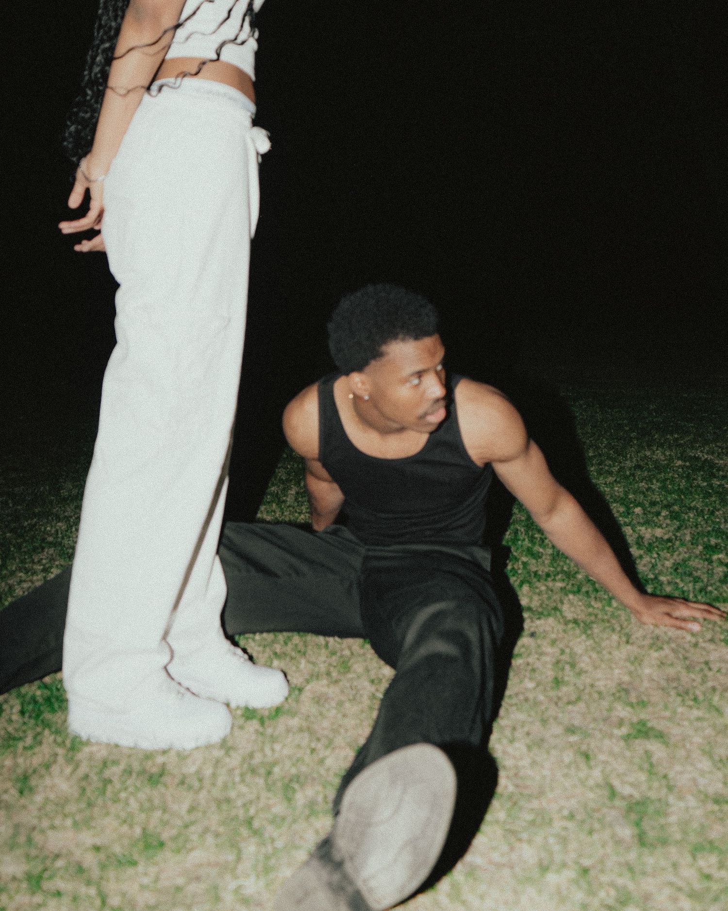 Man in black tank top sitting on grass at night beside person in white pants.