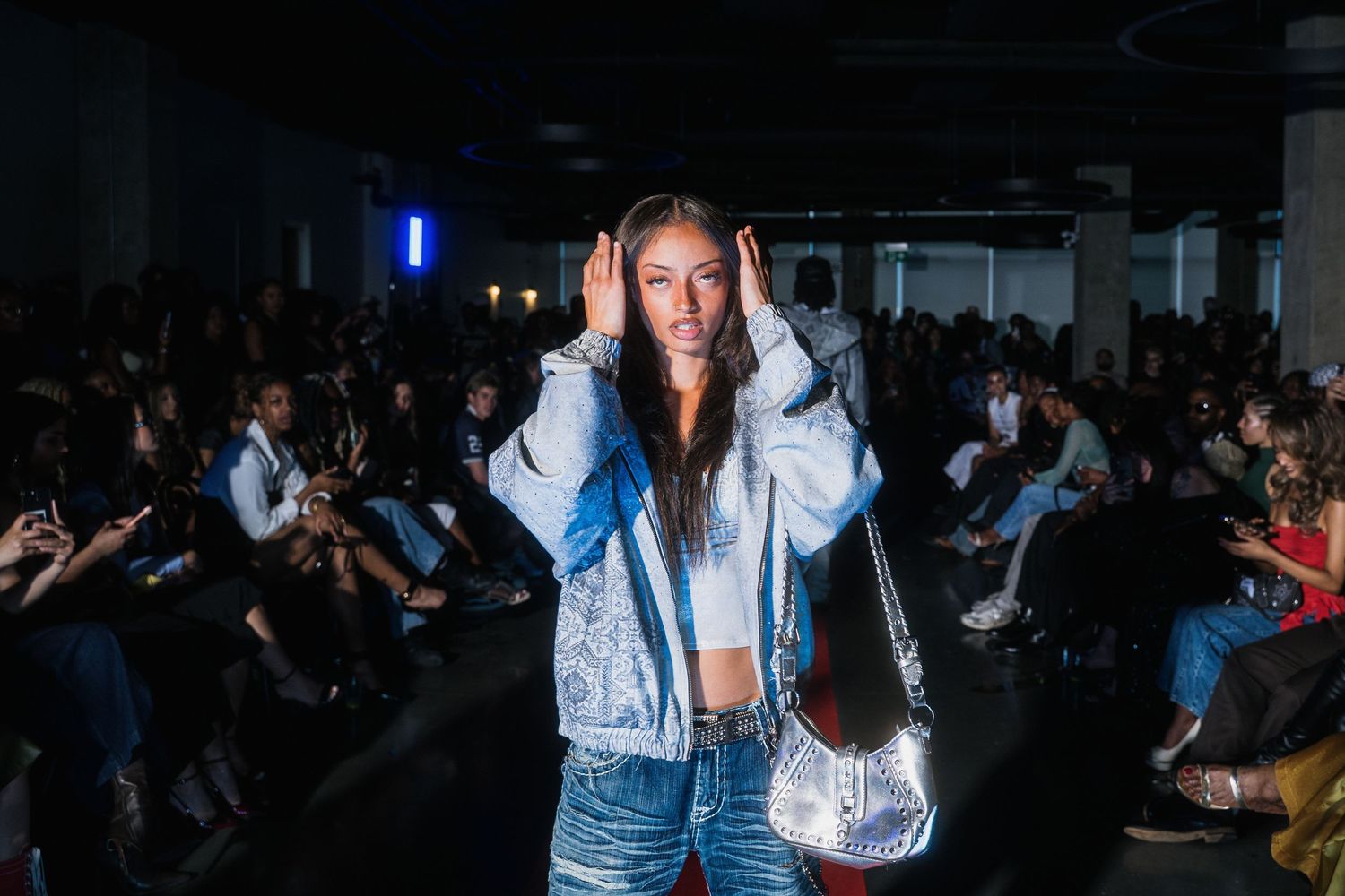 Female model poses on runway in silver denim jacket holding metallic bag at indoor fashion show.