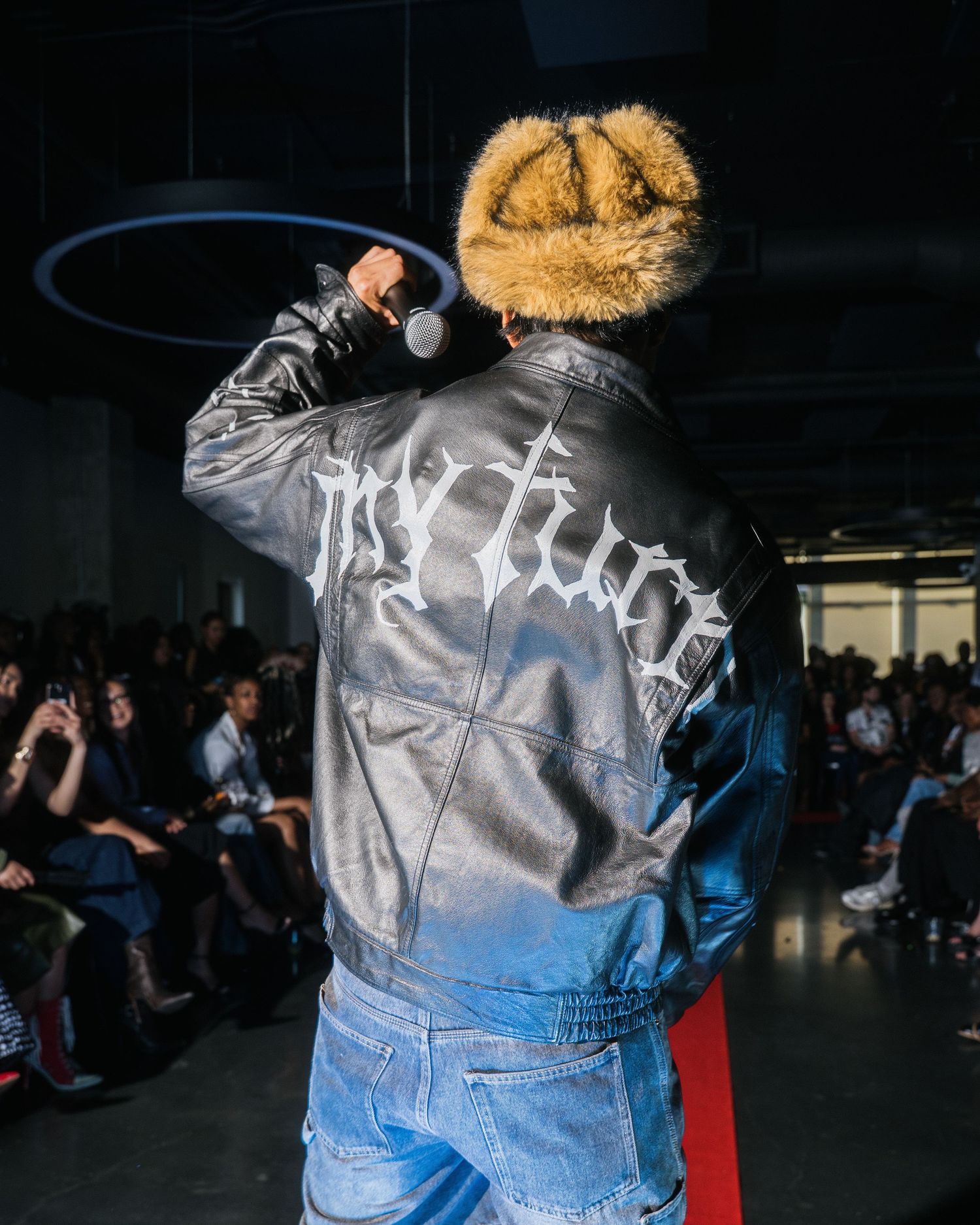 Performer in fur hat and My Turn branded leather jacket holds microphone on fashion runway.