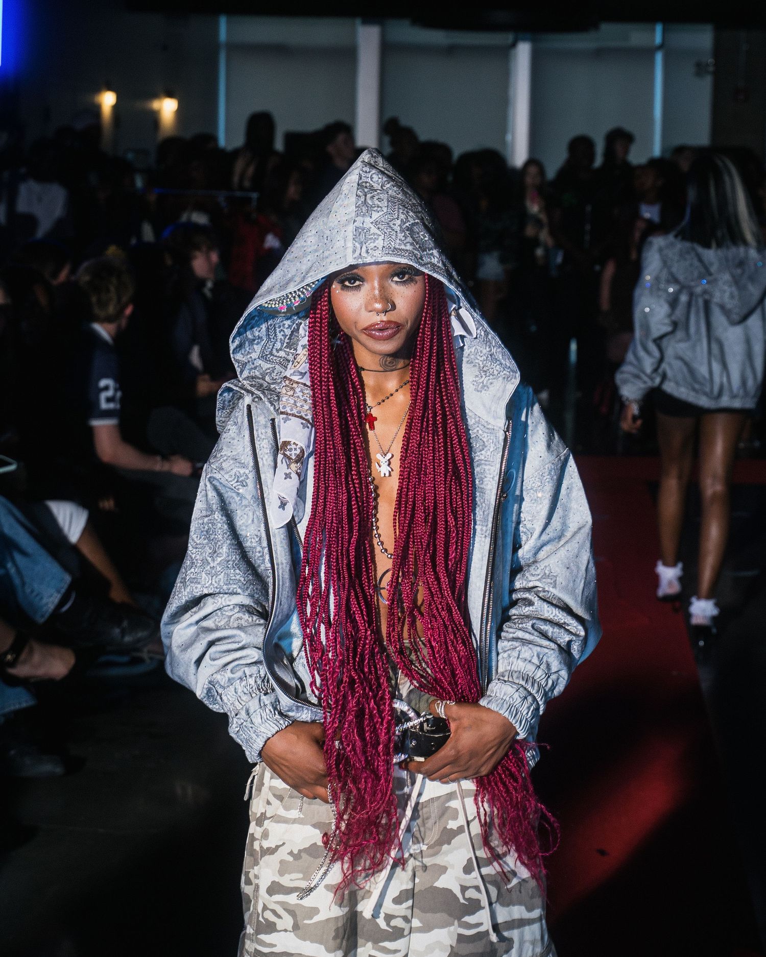 Woman with long red braids wearing a silver hooded jacket and camo pants at a fashion event.