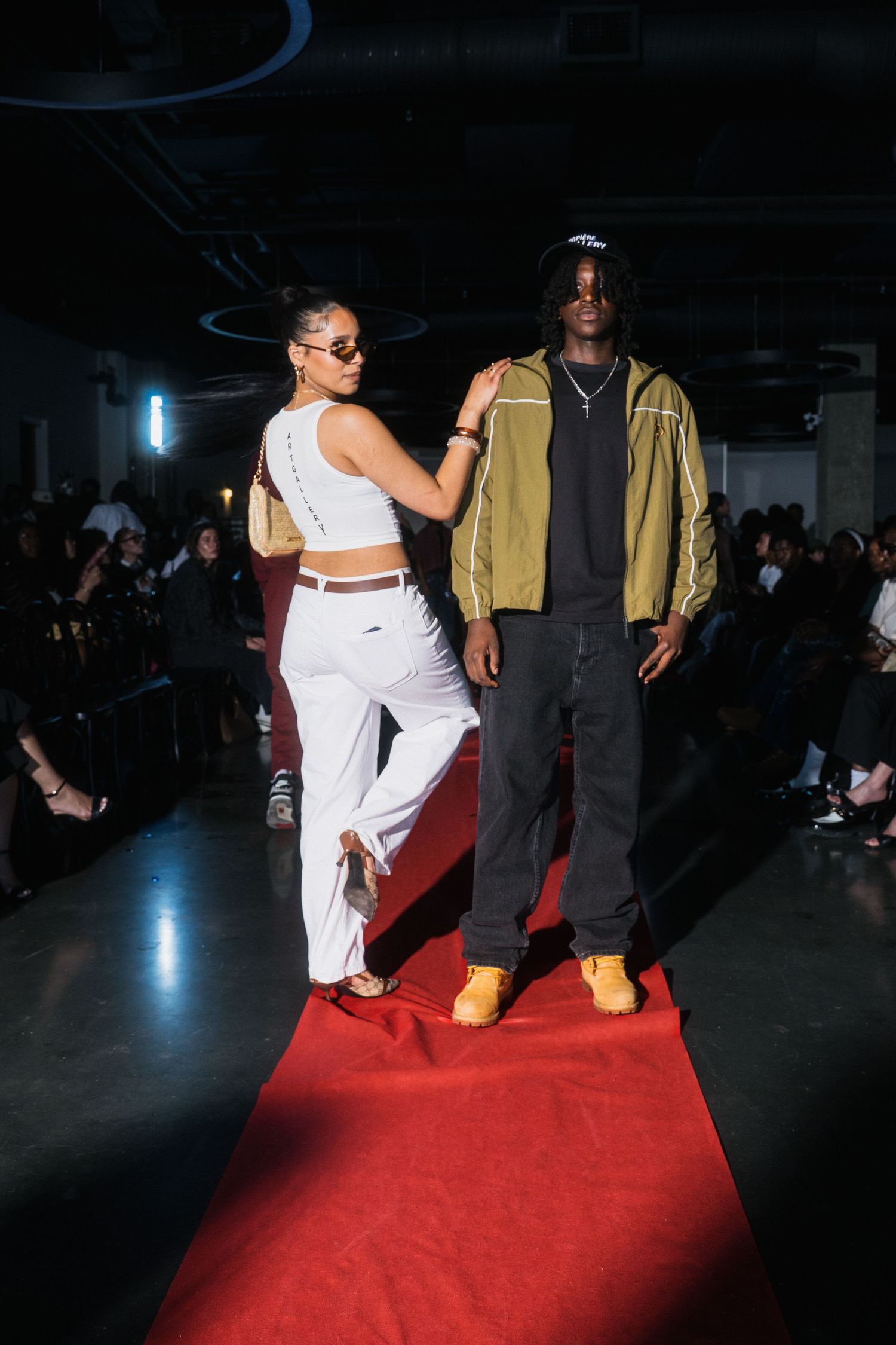 Male and female models pose on red carpet runway, wearing white outfit and olive jacket with yellow boots.