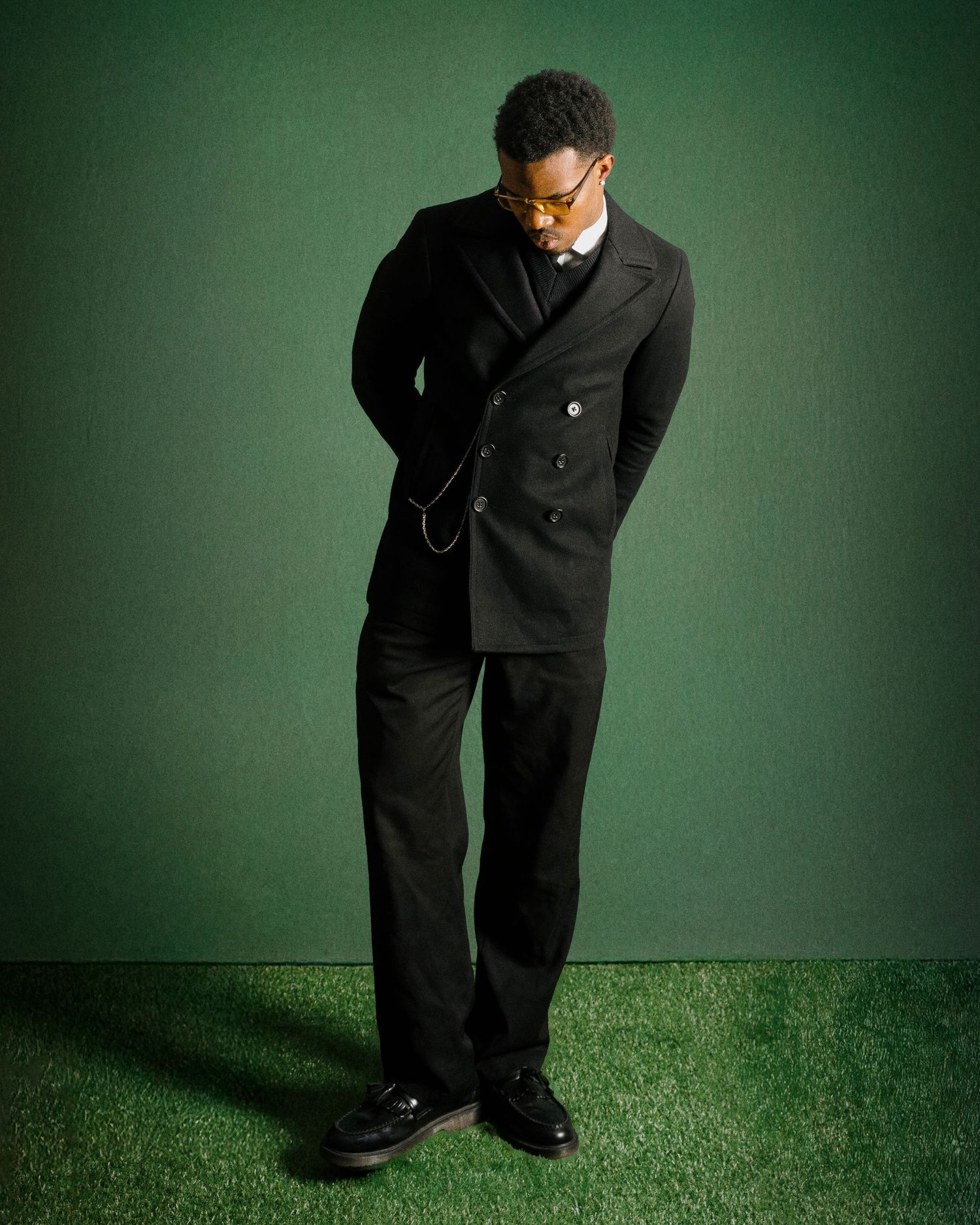 Stylish man in black double-breasted suit looking down on green grass backdrop.