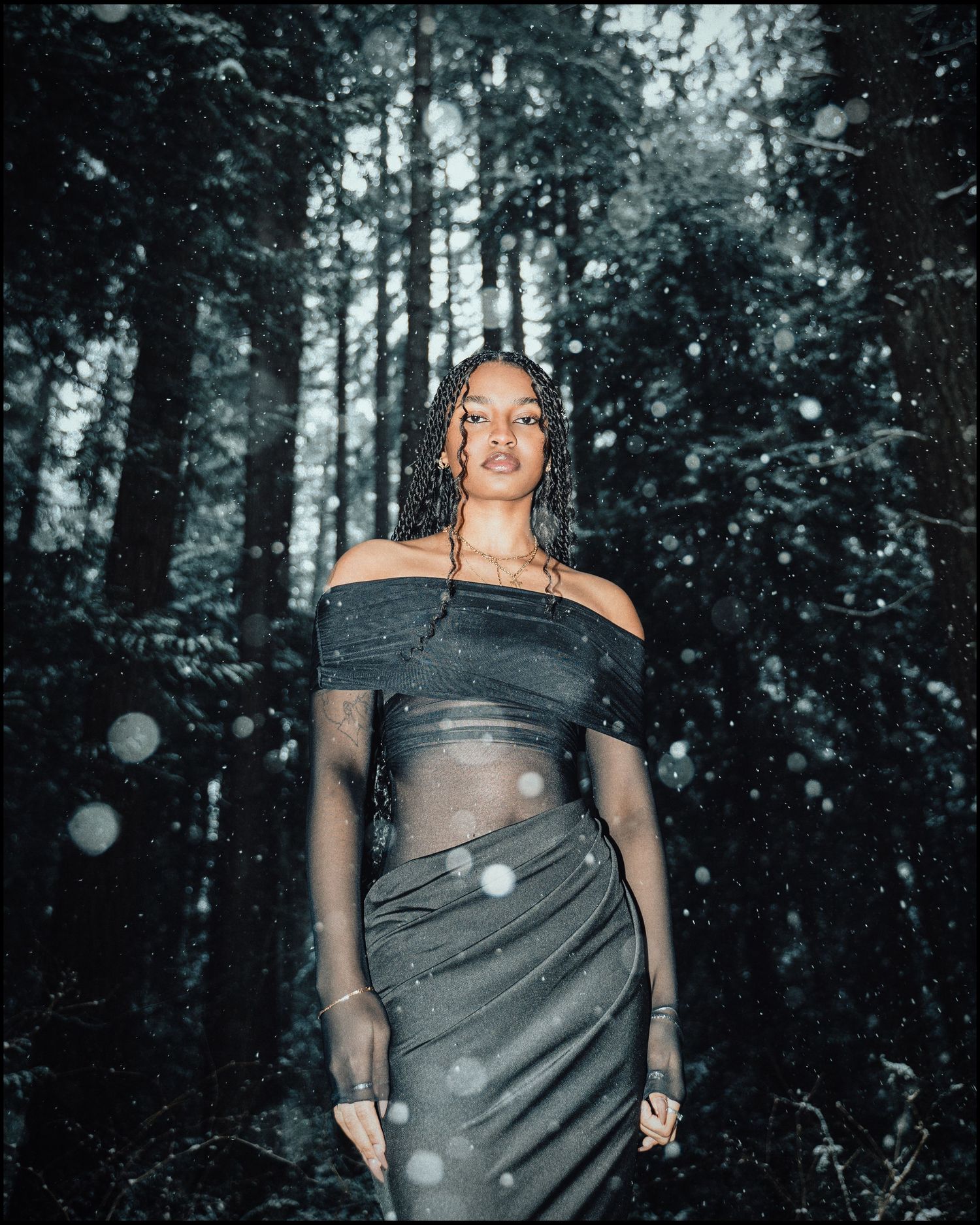 Woman in grey off-shoulder outfit standing in dark snowy forest at night.