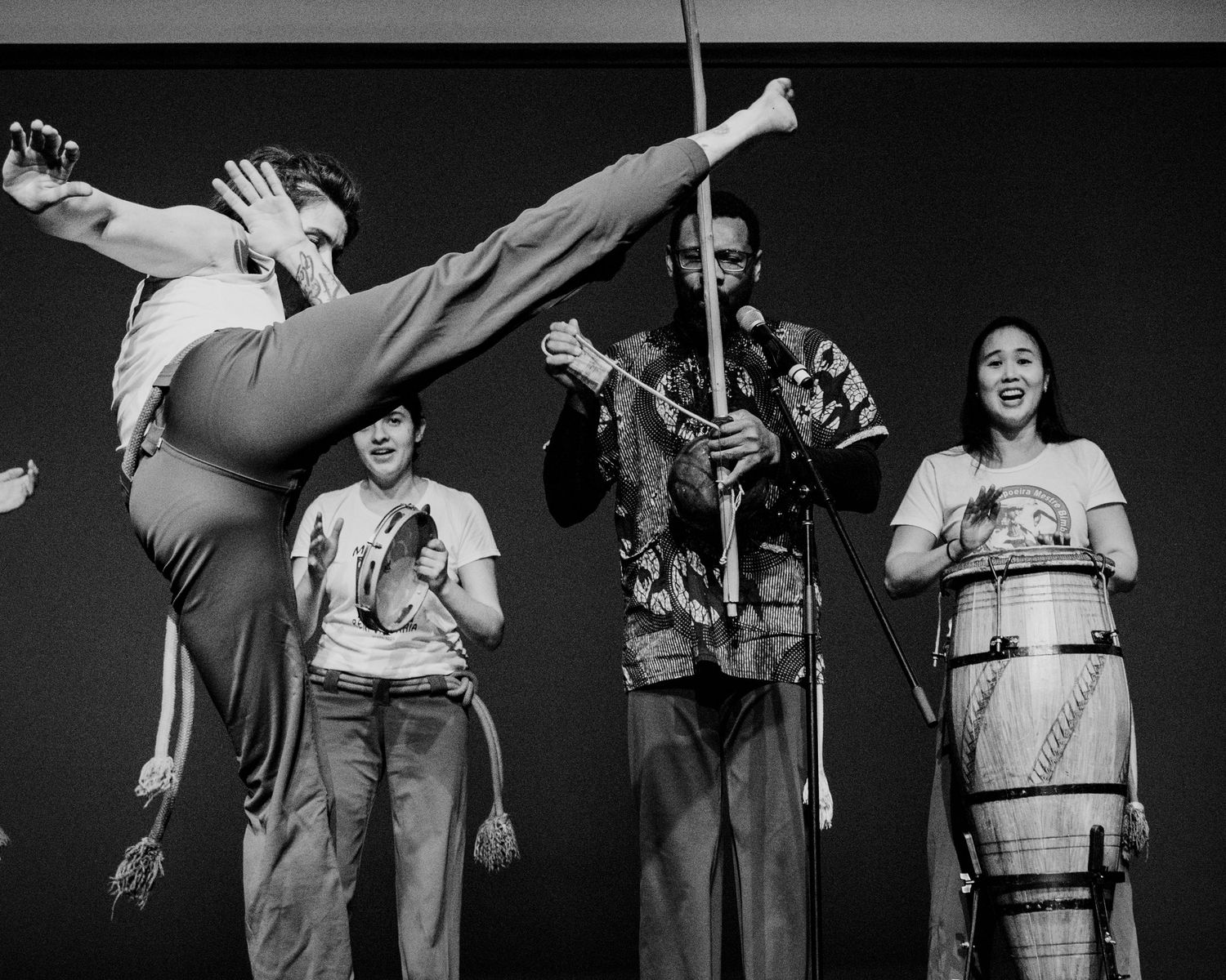 Capoeira dancer executes high kick on stage alongside musicians with drums and staff.
