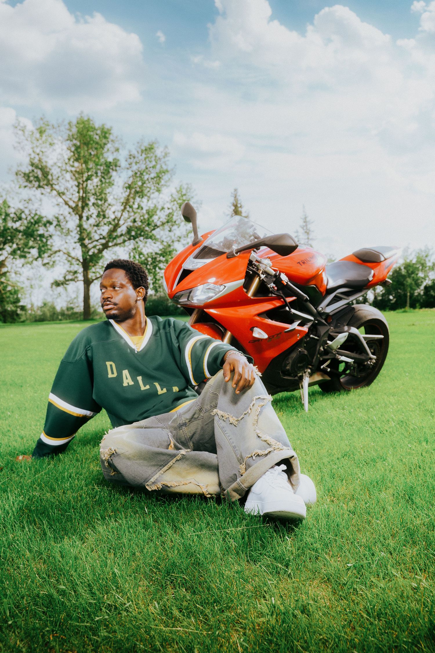 Man in Dallas green jersey and distressed jeans sitting on grass beside a red sport motorcycle outdoors.