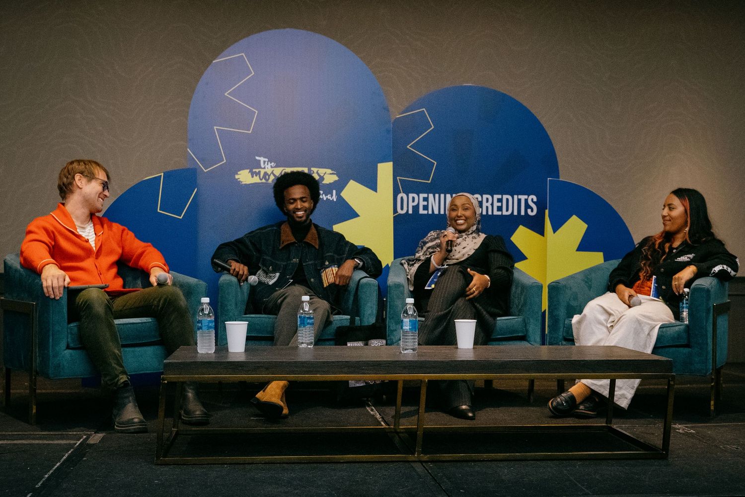 Four panelists sit on teal chairs during a discussion event in front of a blue branded backdrop.