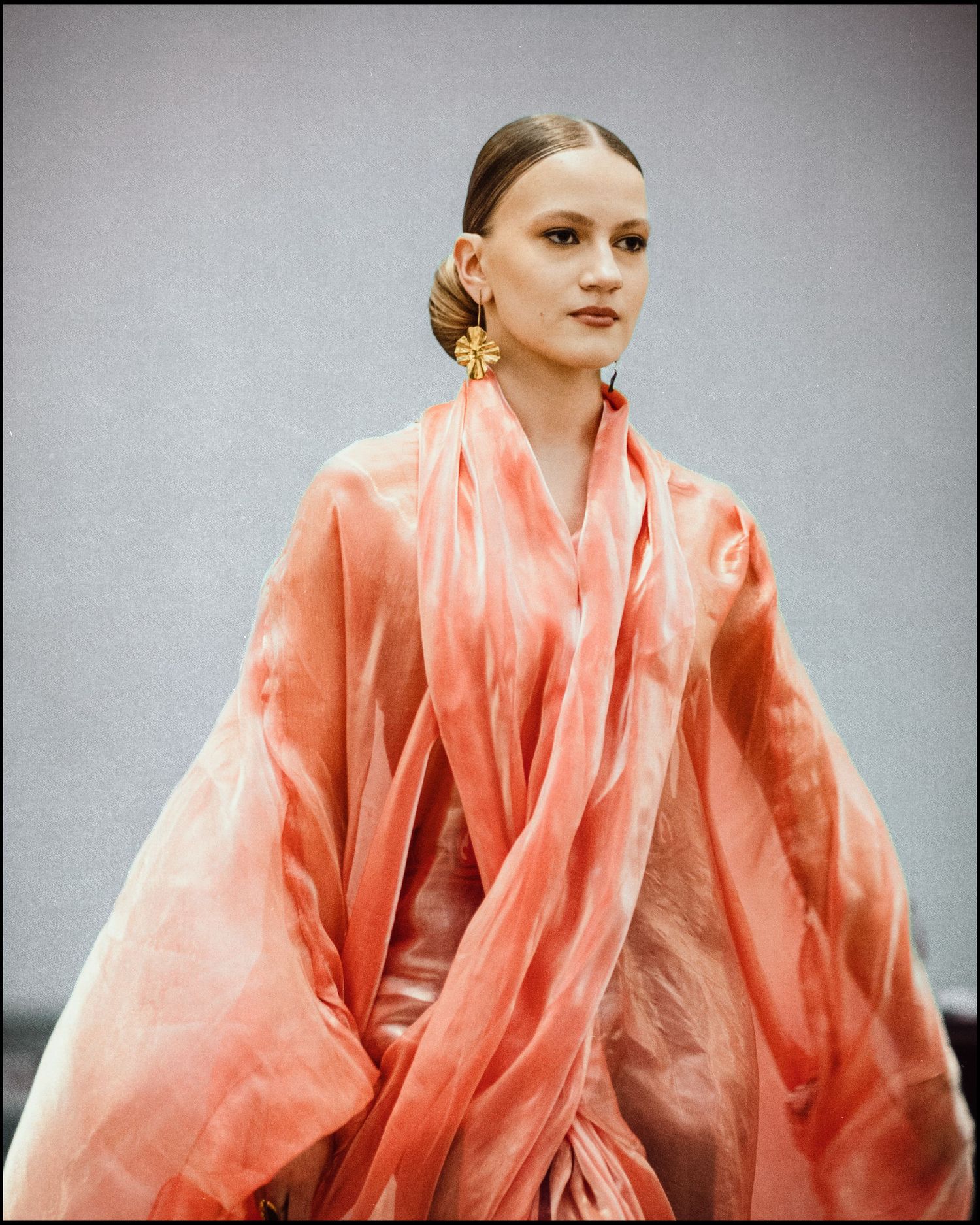 Close-up of model wearing flowing coral pink sheer organza wrap with large gold floral earrings and sleek updo.
