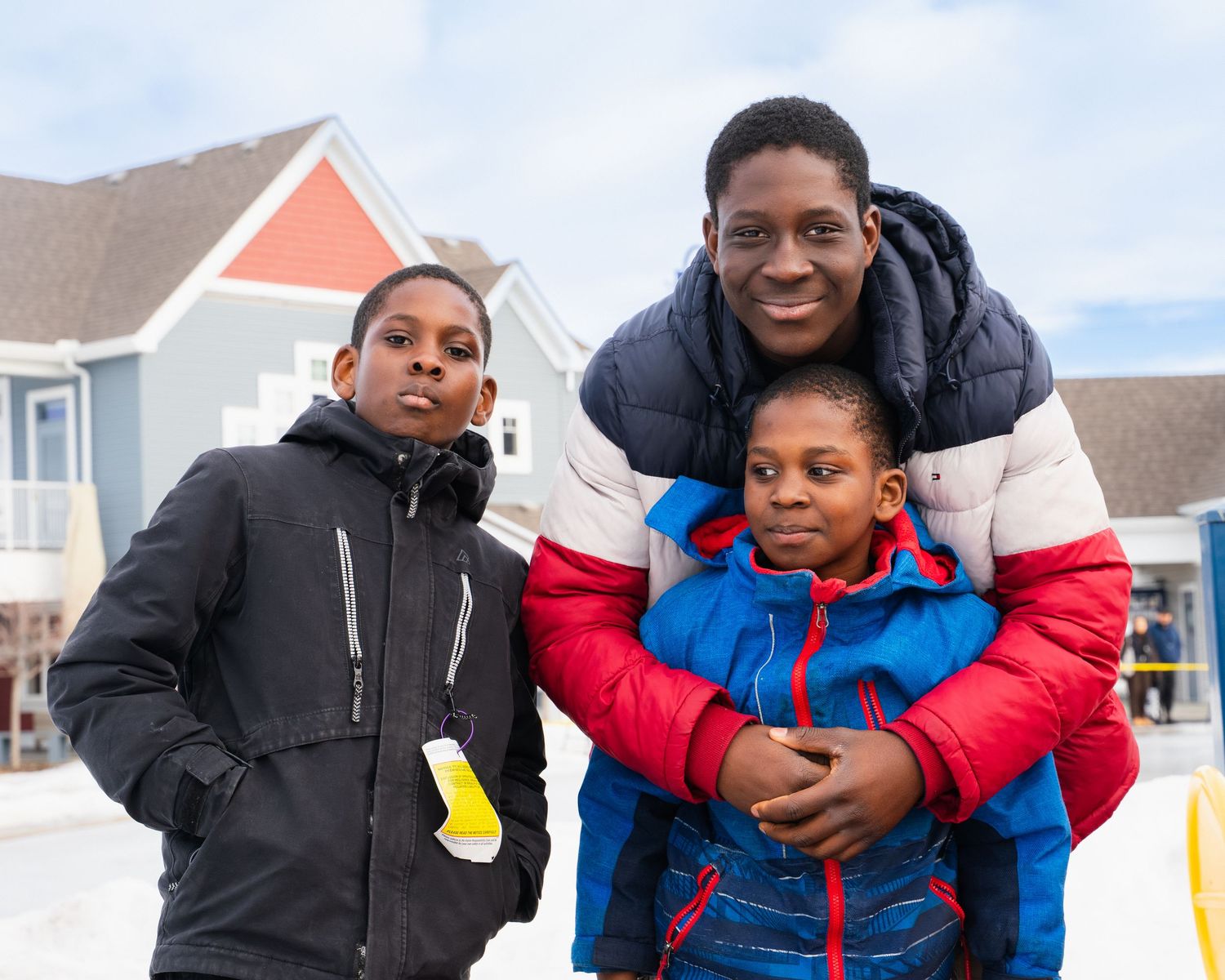 Three young boys in winter jackets posing together outdoors in snowy residential neighborhood.