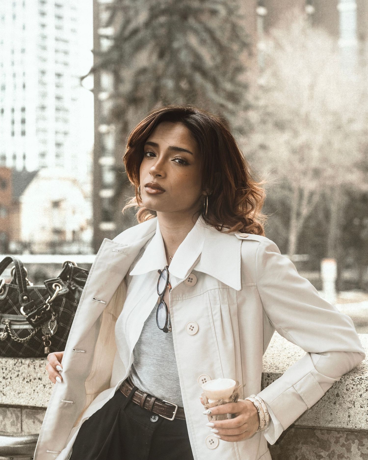 Stylish woman in cream trench coat holding coffee cup in urban city setting.