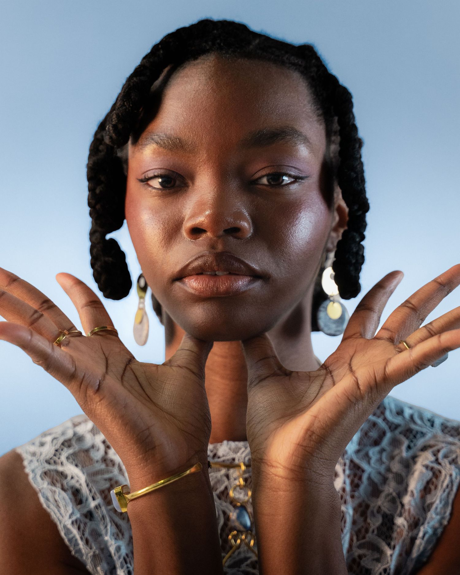 Close-up portrait of a Black woman with braids displaying jewelry and rings against blue background.
