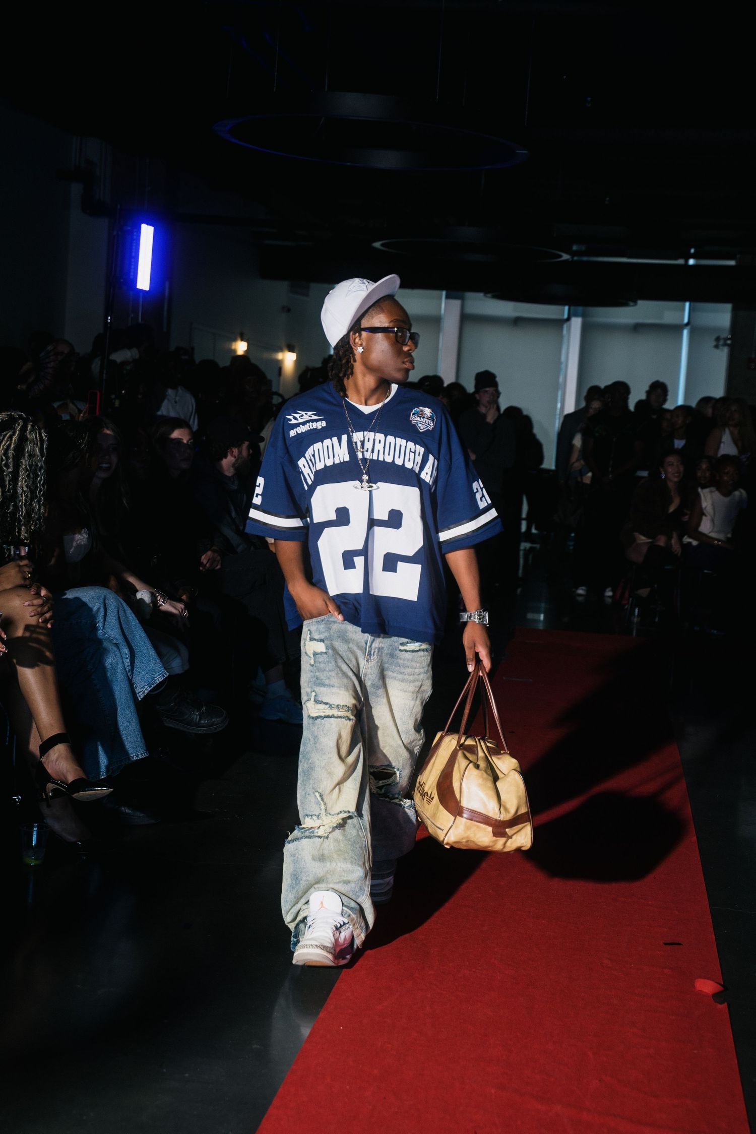 Male model walks red carpet in blue number 22 jersey, baggy jeans, and cap at fashion show.