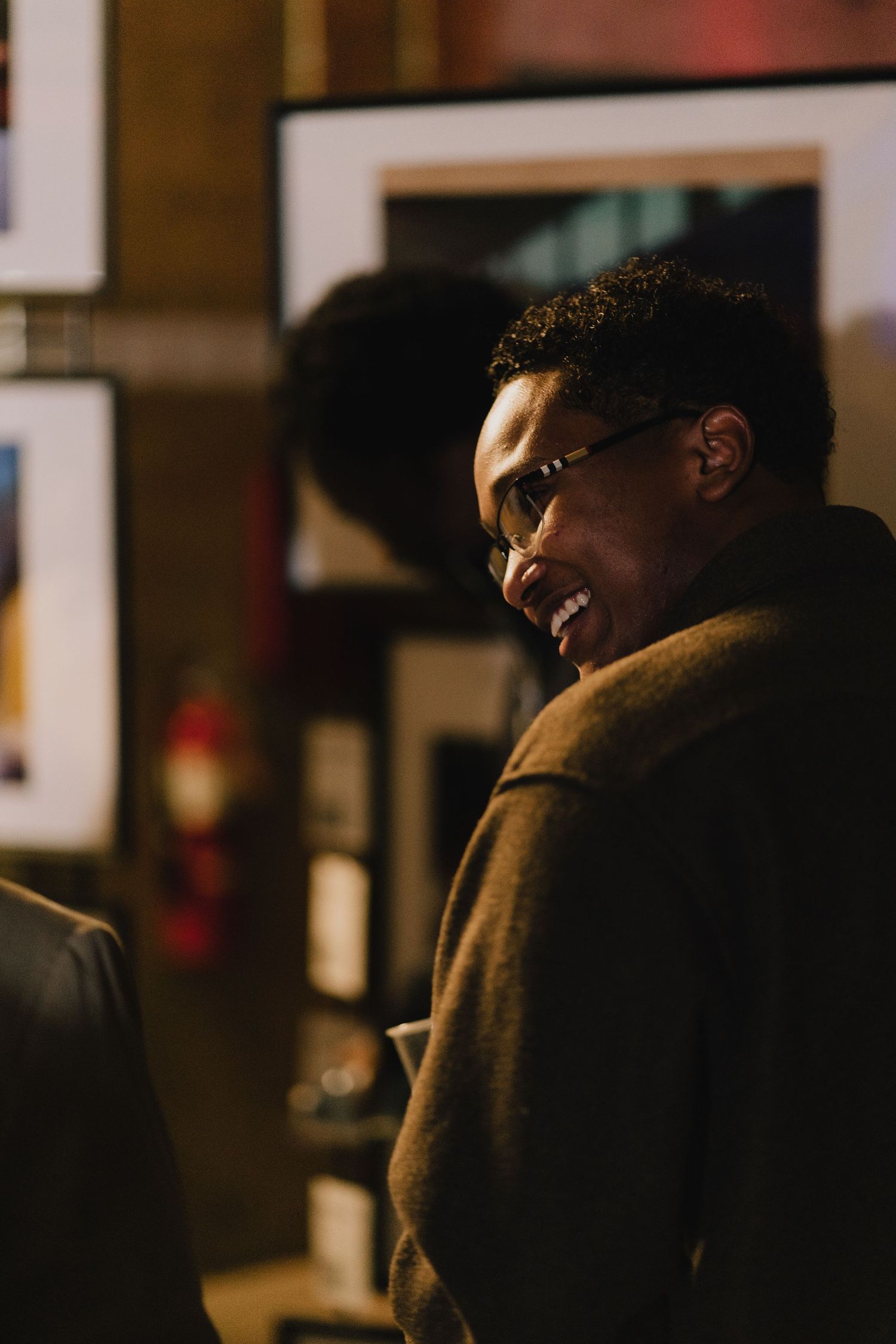 A smiling man with glasses and a dark coat looks back warmly in a dimly lit gallery setting.