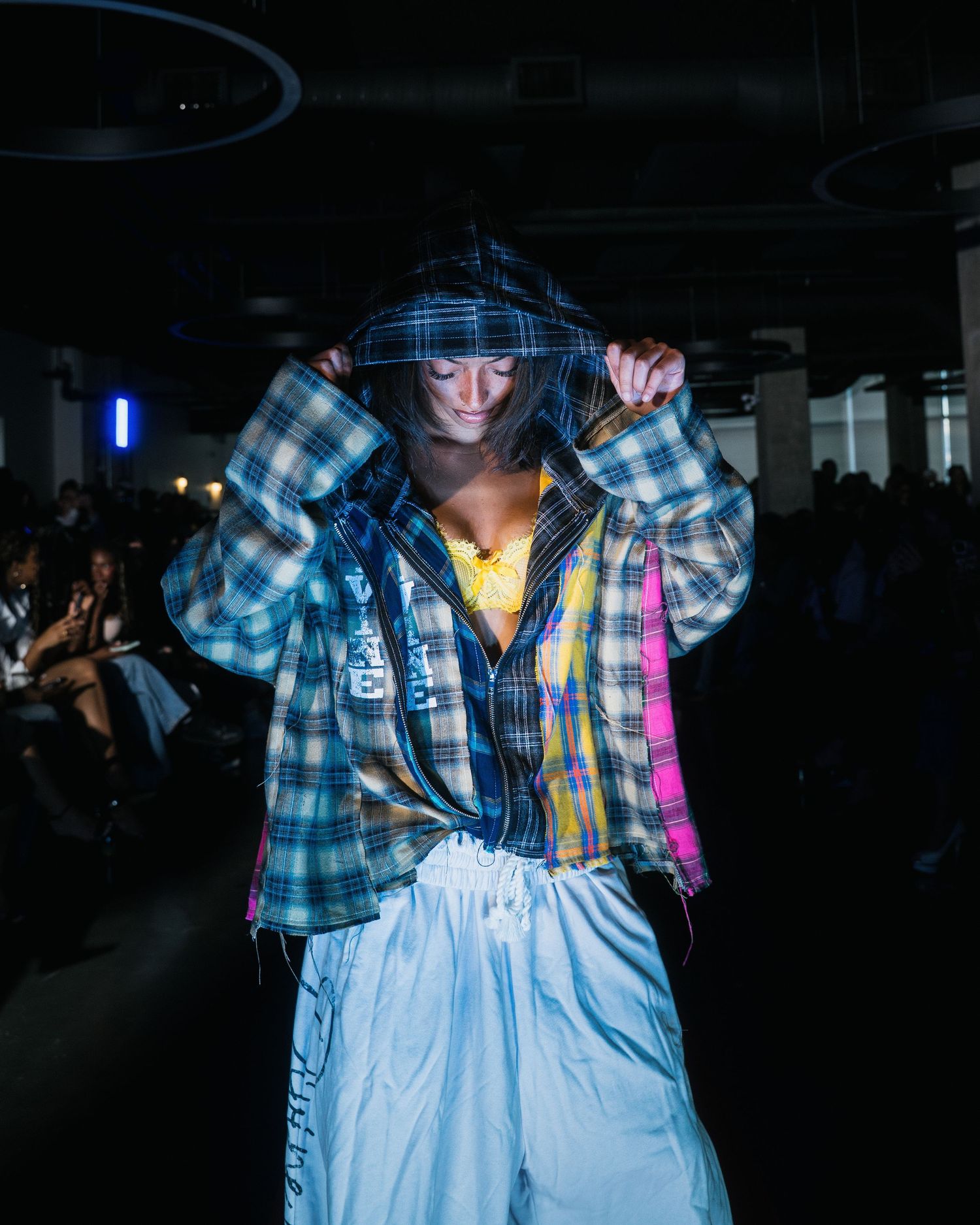 Model walks runway in patchwork plaid hoodie and white cargo pants at an indoor fashion show.