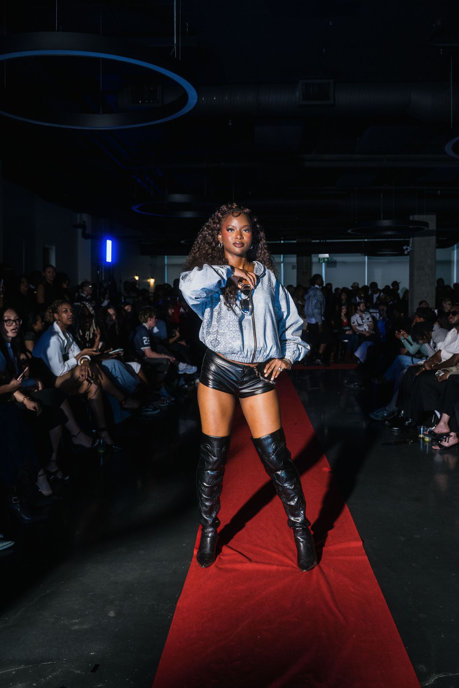Female model poses on red carpet runway in silver jacket and black leather shorts with thigh-high boots.