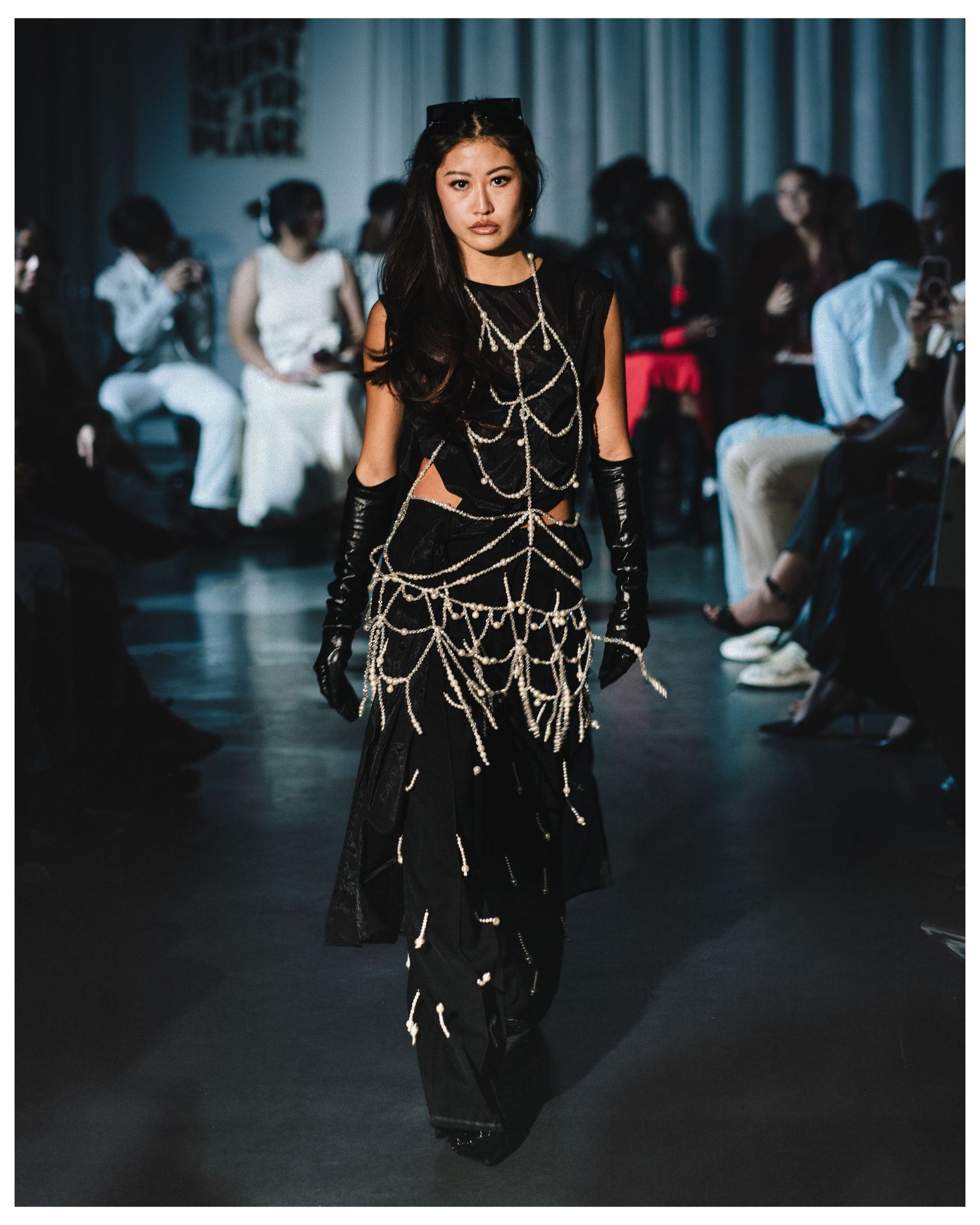 Model on runway wearing black outfit with gold chain details and long black gloves.