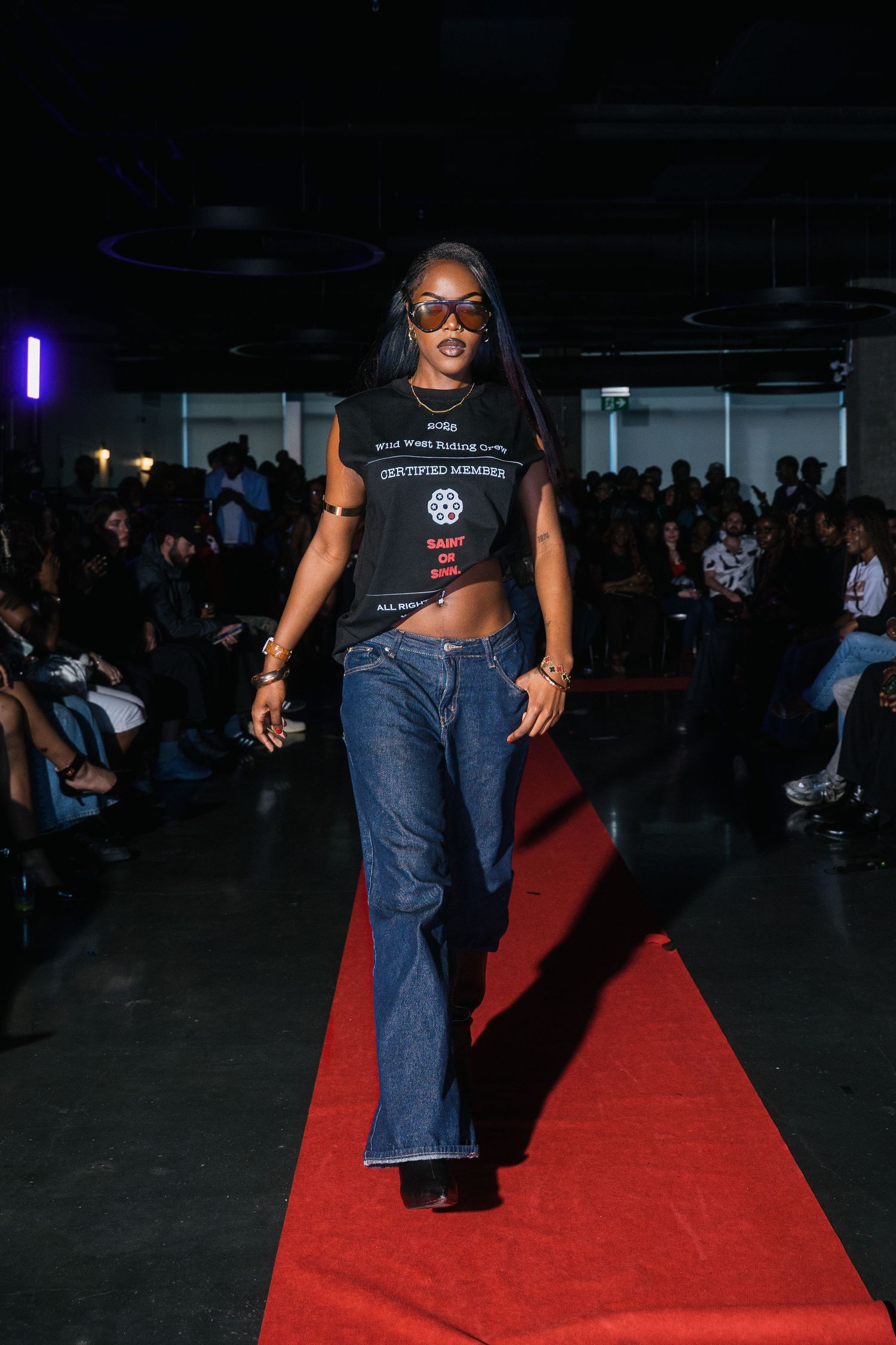Female model struts runway in black graphic crop tee, wide-leg jeans, and oversized sunglasses.