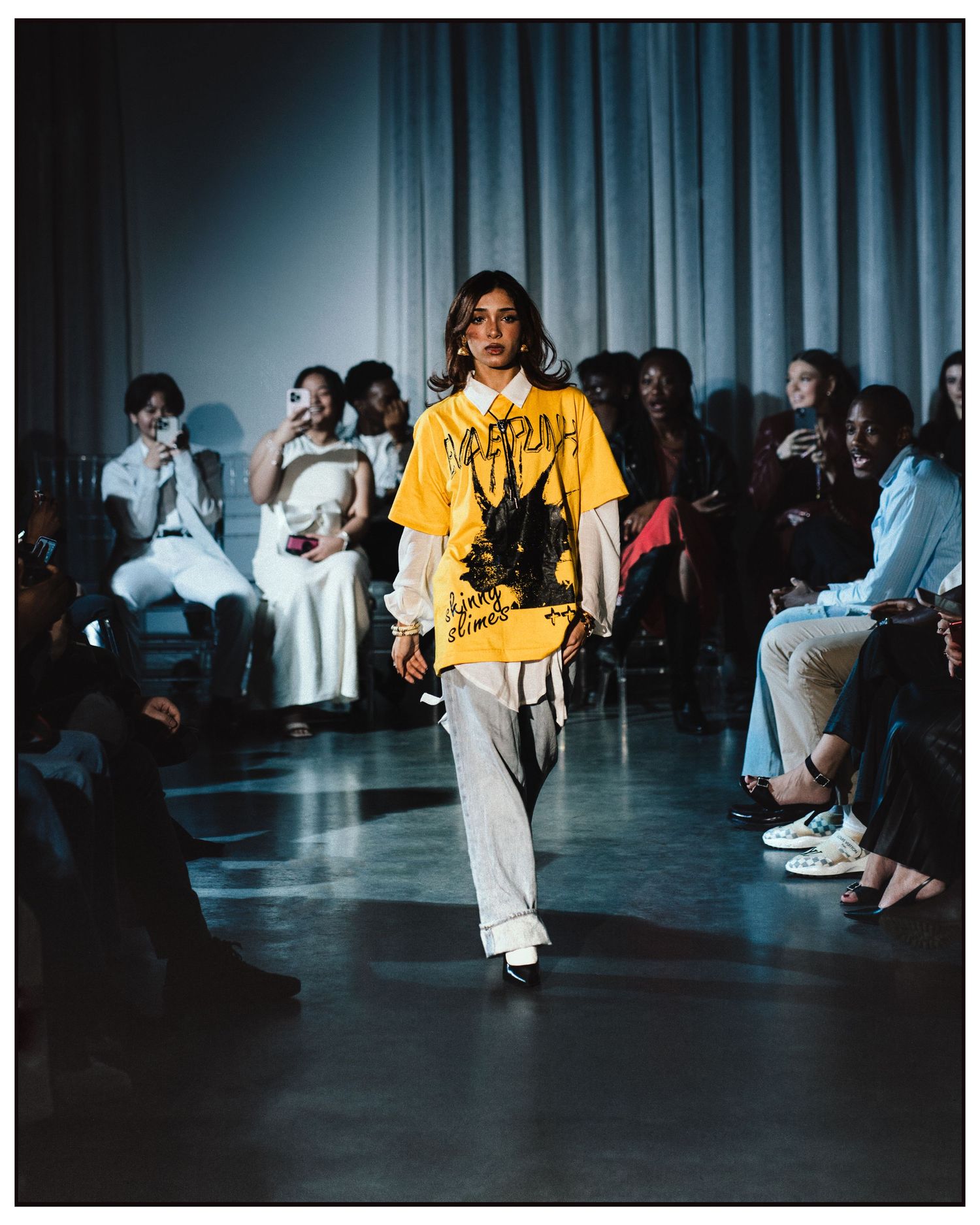 Model walks runway in oversized yellow graphic tee and white trousers at fashion show.