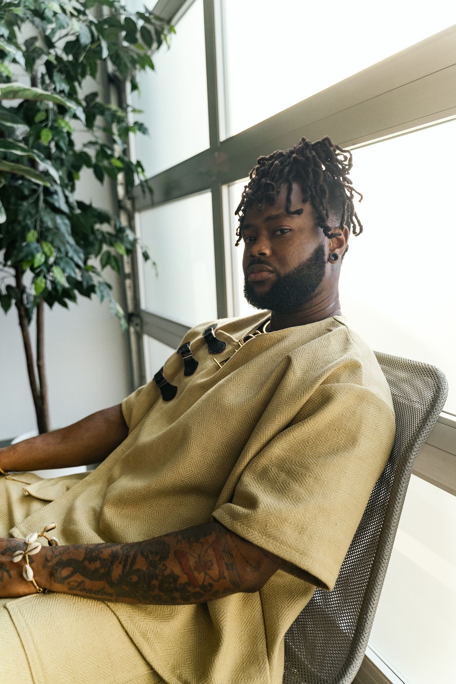 A young Black man with dreadlocks and tattoos sits relaxed in a mesh chair wearing a beige outfit.