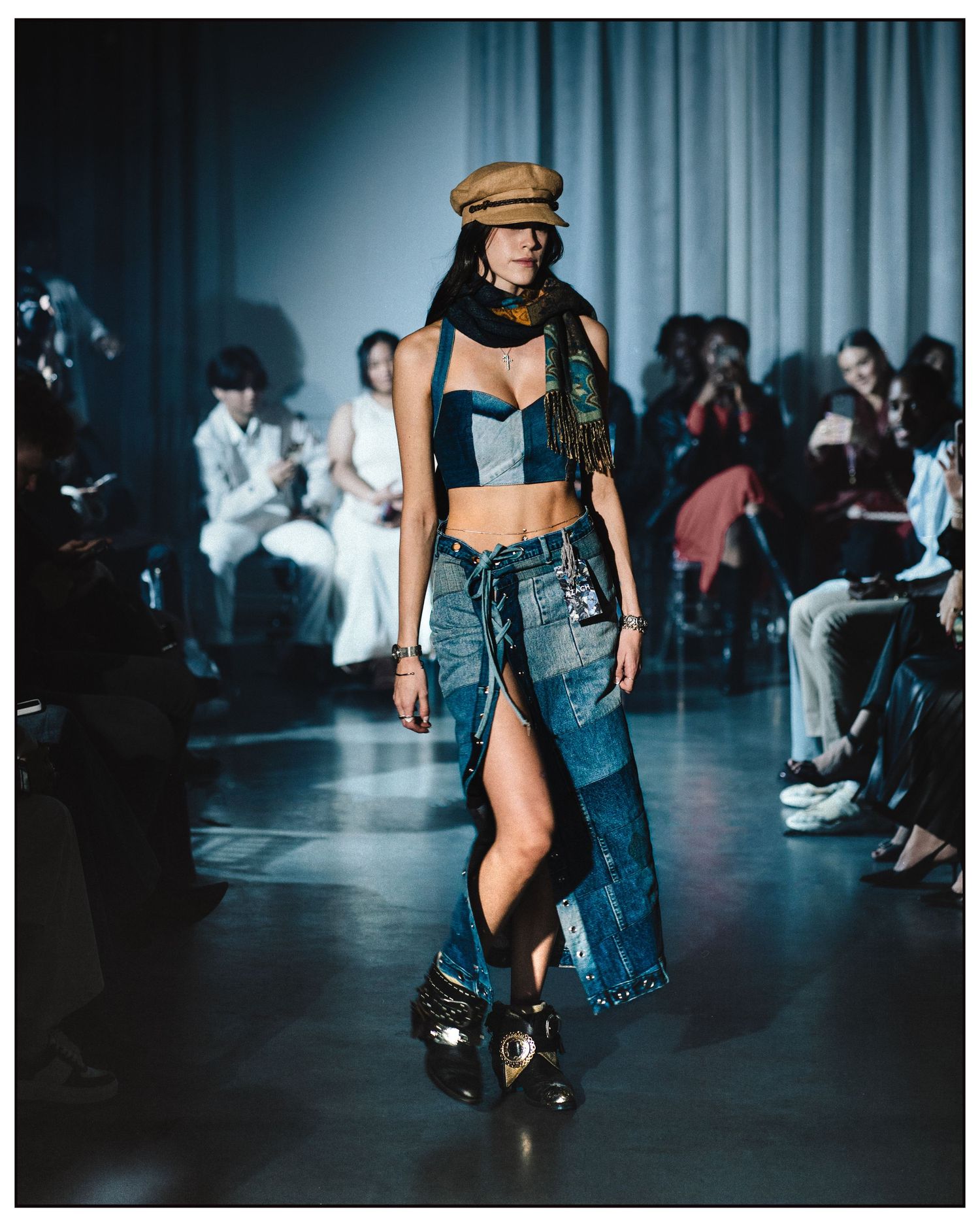 Model walks runway in denim bralette, patchwork maxi skirt, and newsboy cap at fashion show.