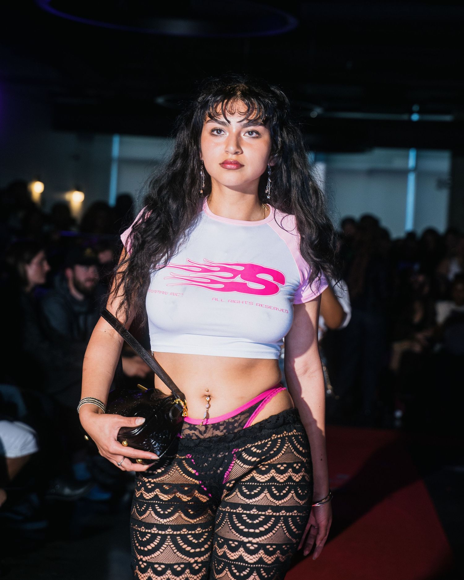 Female model poses on runway in white crop top with pink flame logo and black lace pants.