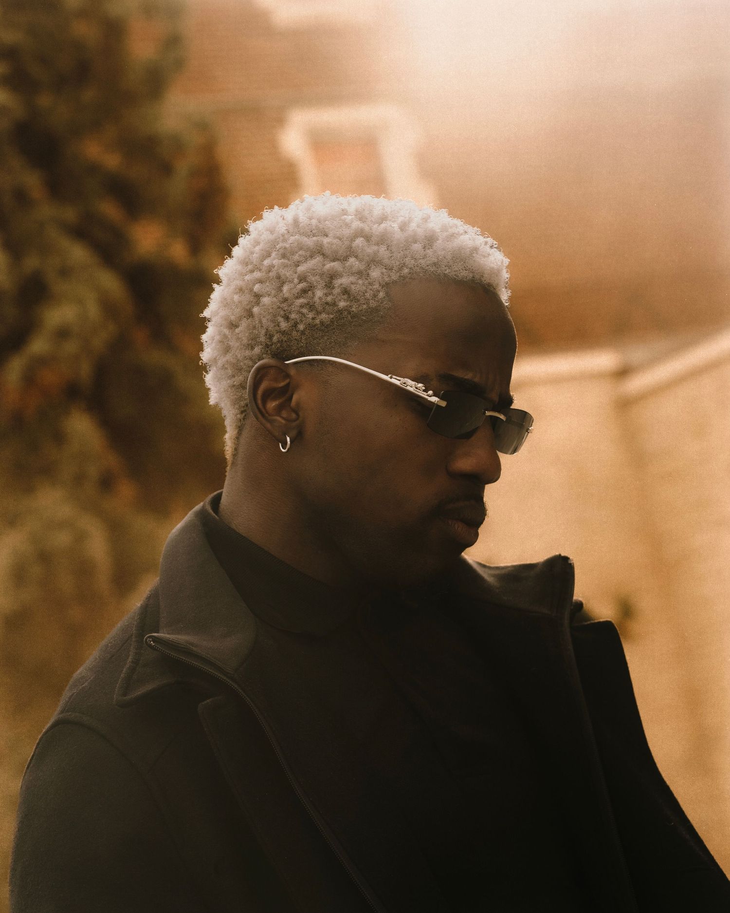 Man with bleached curly hair and sunglasses in black outfit poses outdoors in golden light.