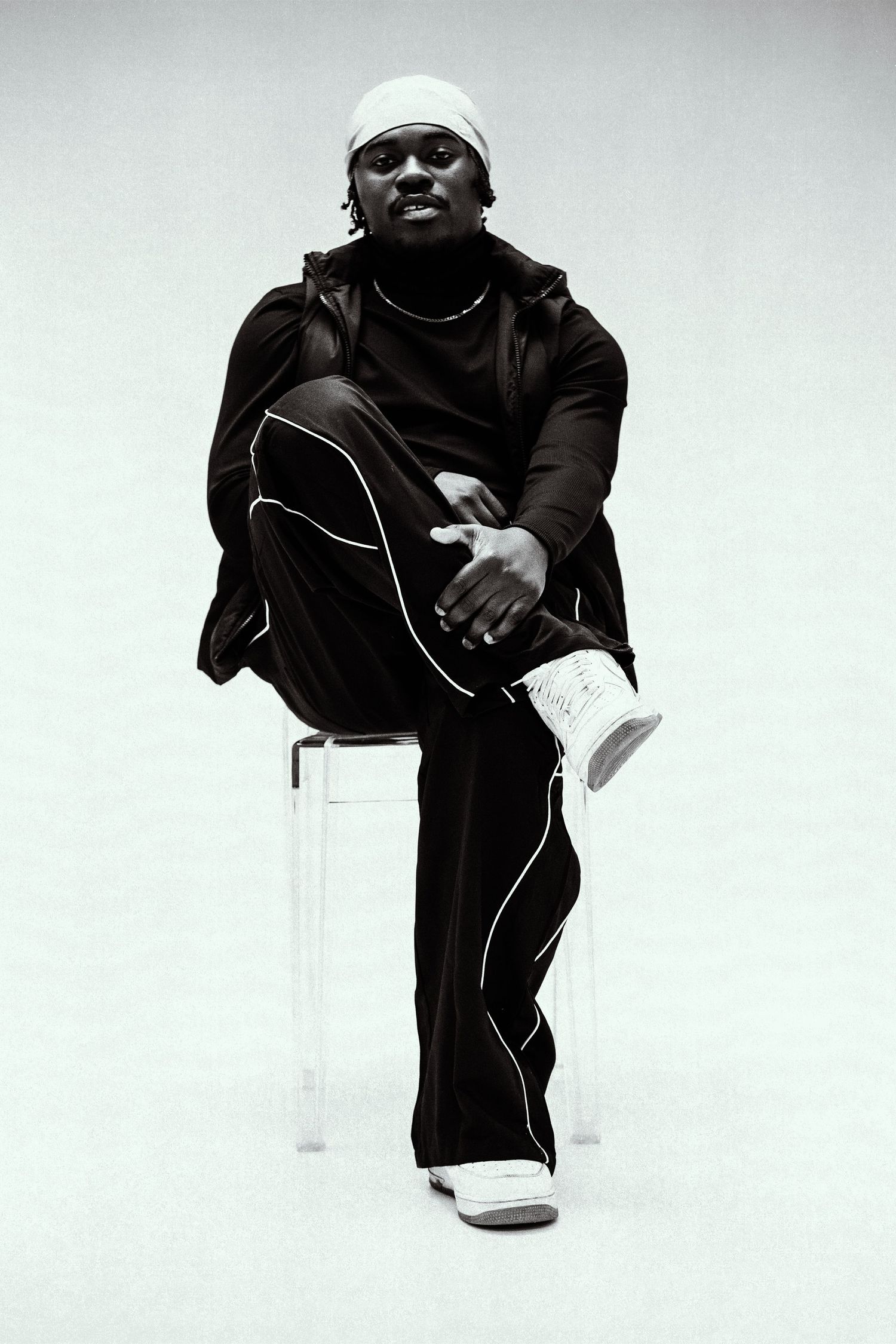 Man in black tracksuit seated on clear chair posing in black and white portrait.