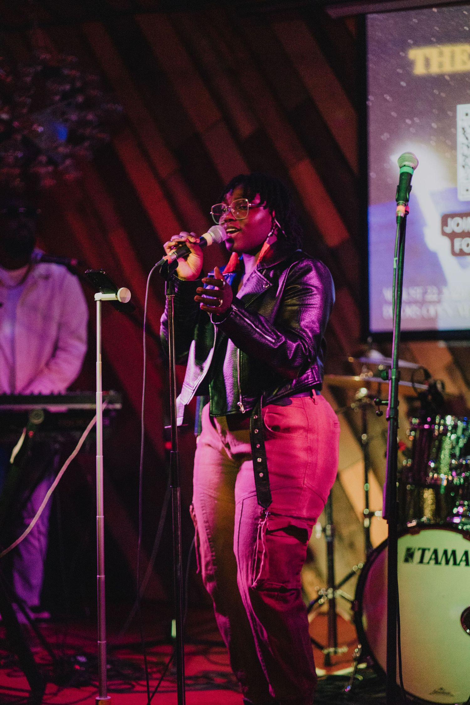 A Black male vocalist performs on stage in a leather jacket under pink stage lights with a Tama drum kit nearby.