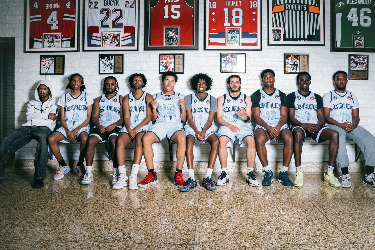 A basketball team poses together on a bench in front of framed jerseys on a brick wall.