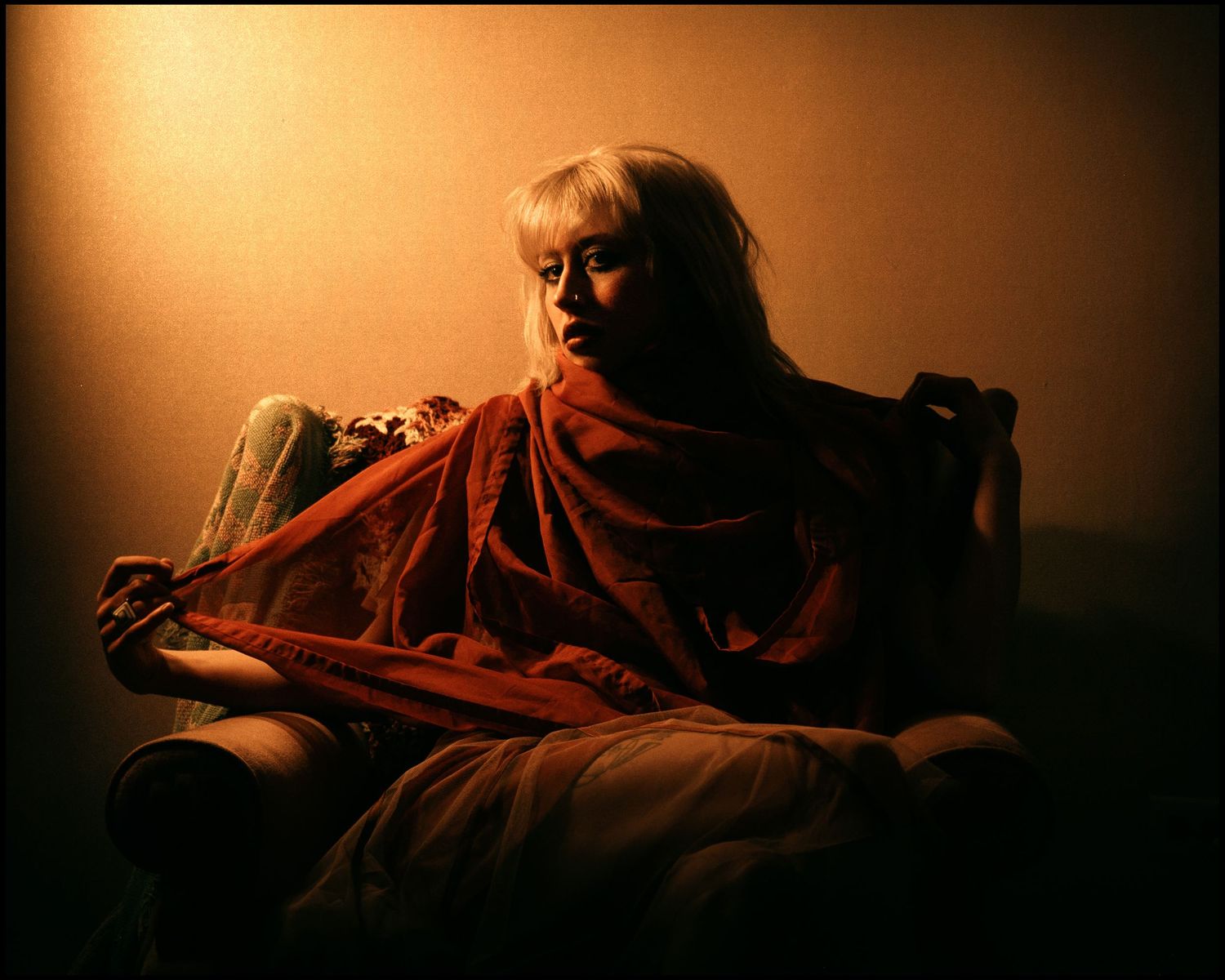 A blonde woman wrapped in a red shawl sits in a chair bathed in warm golden ambient light.