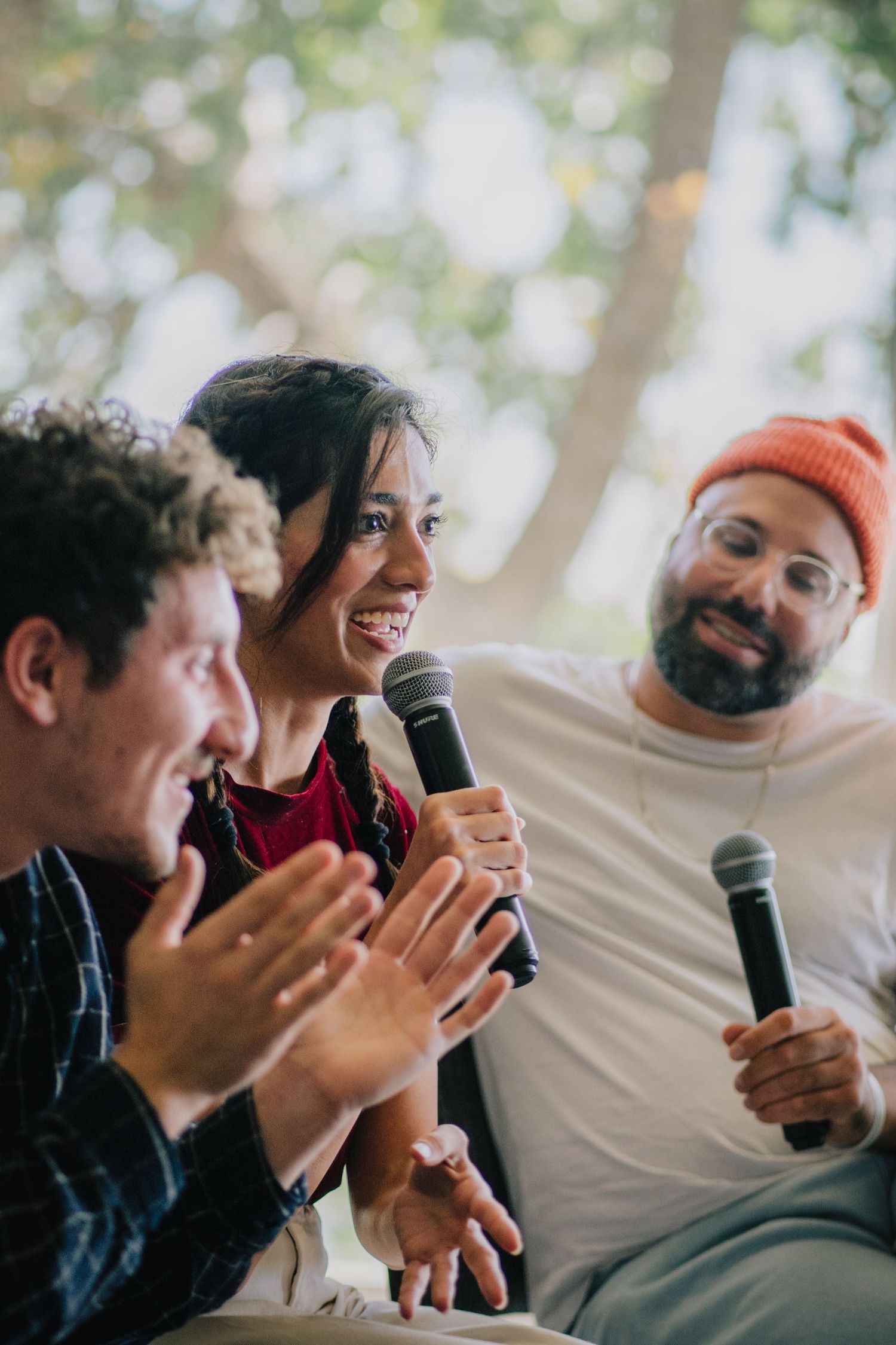 Three diverse panelists smile and engage during a lively discussion, with two holding microphones at an outdoor event.