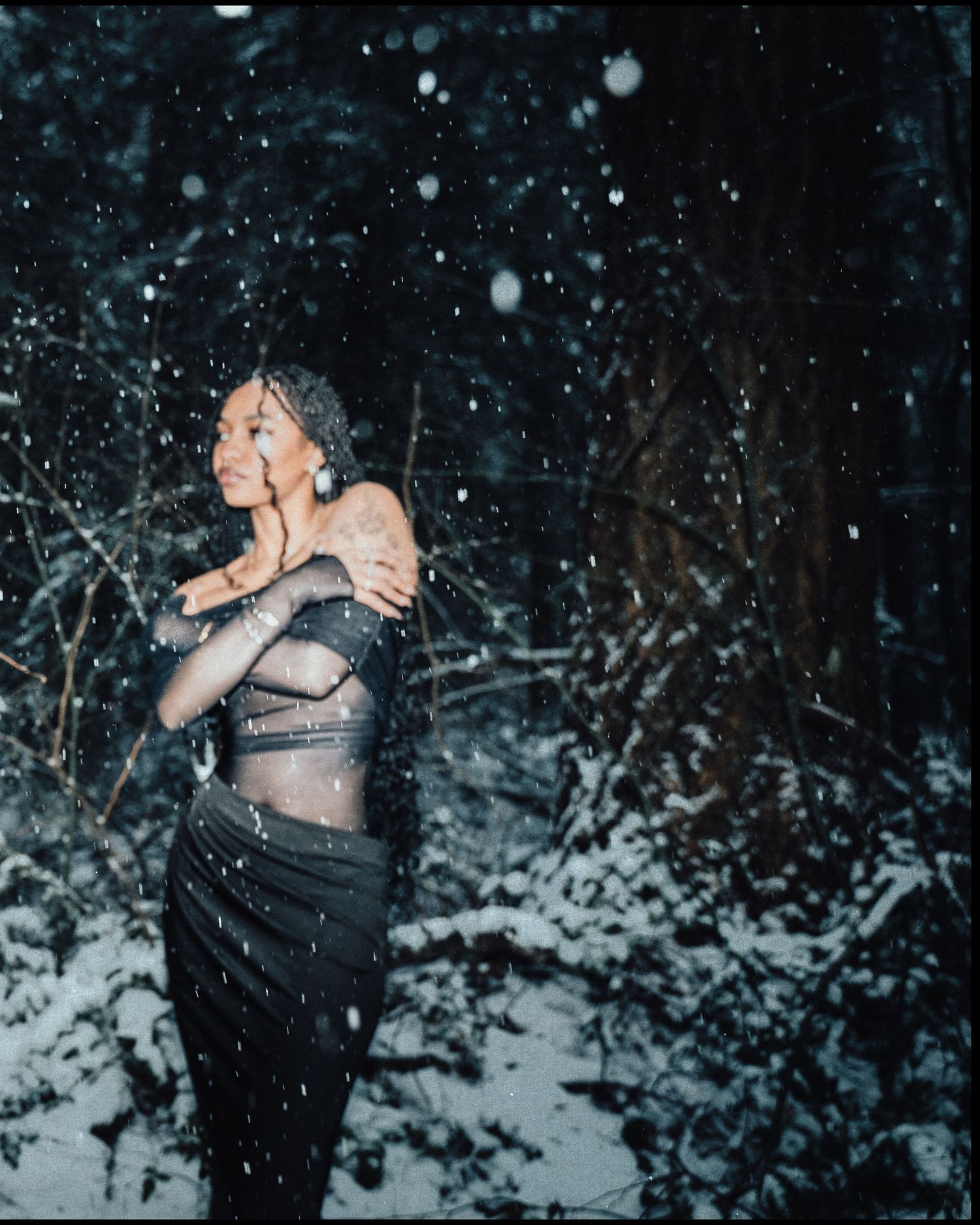Woman in sheer grey outfit poses gracefully in a snowy nighttime forest setting.