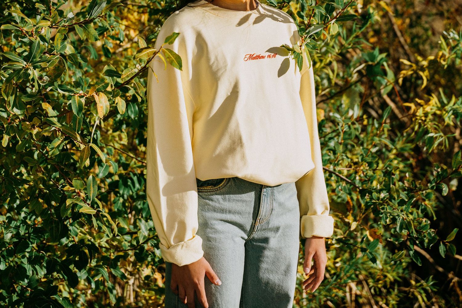 Woman in cream sweatshirt and jeans standing in front of green leafy bushes outdoors.