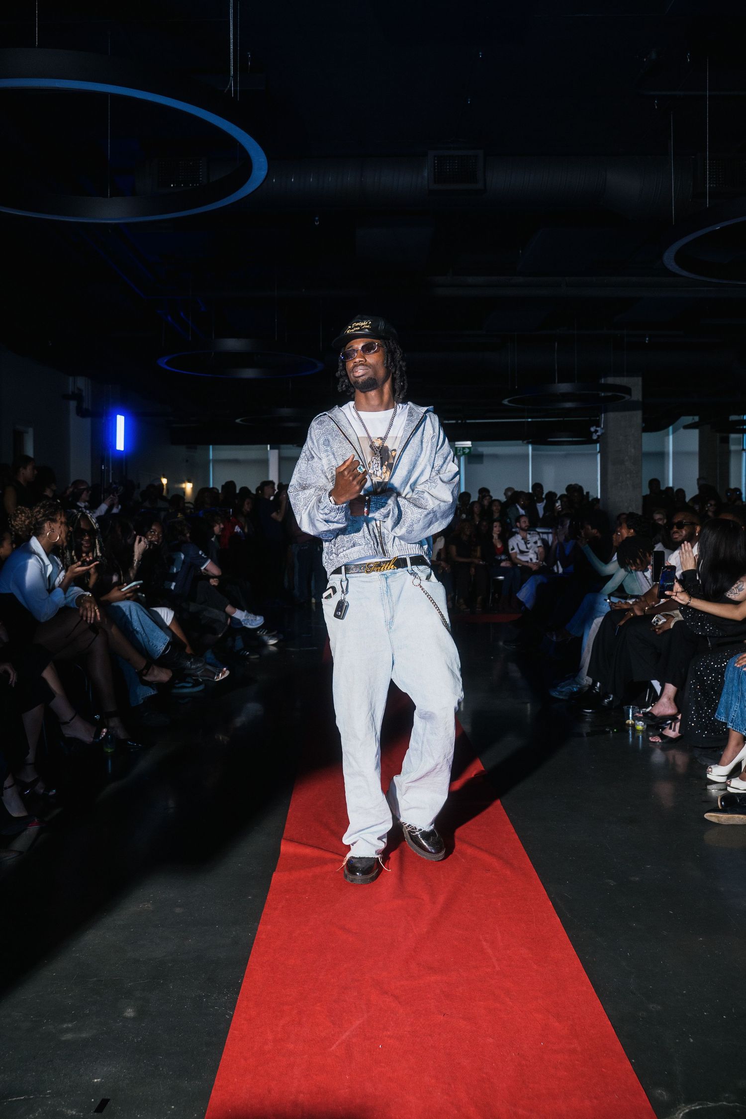 Male model walks red carpet runway in all-white outfit with silver patterned jacket at fashion show.