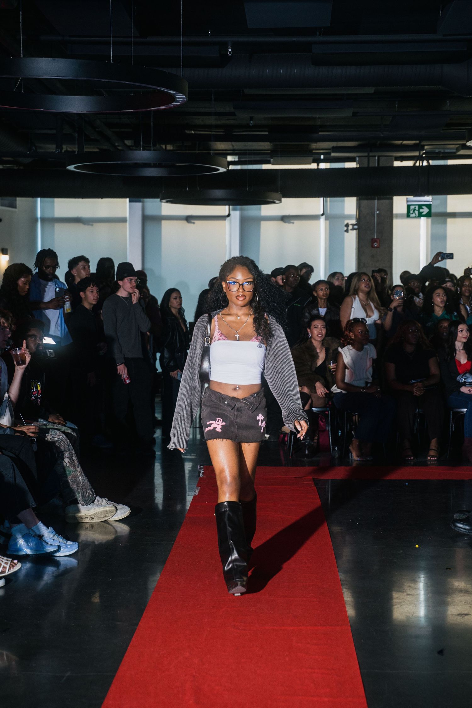 Female model walks red carpet runway in gray cardigan and dark mini skirt with boots.