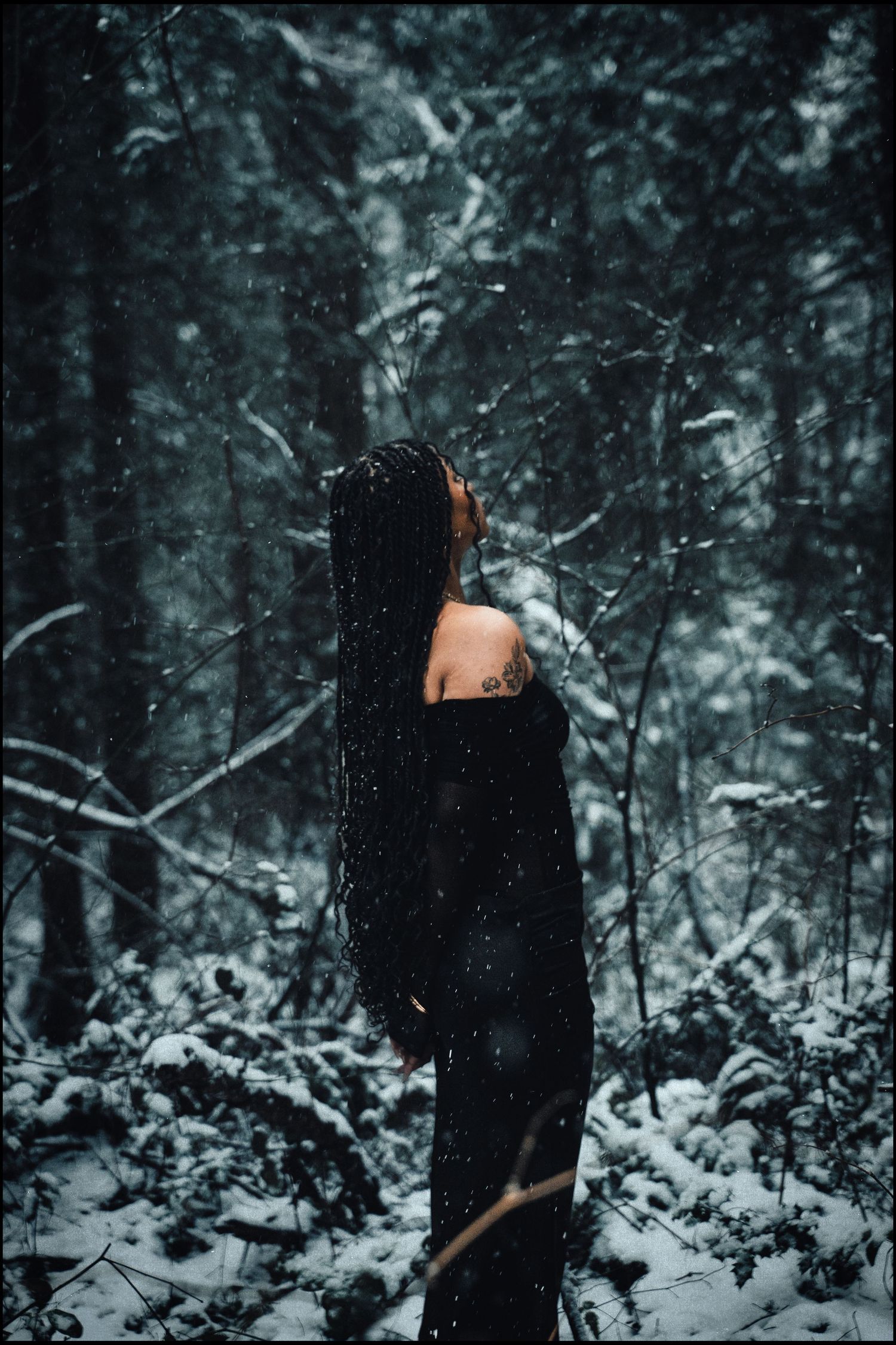 Woman in black dress standing in snowy forest with braids, snow falling around her.