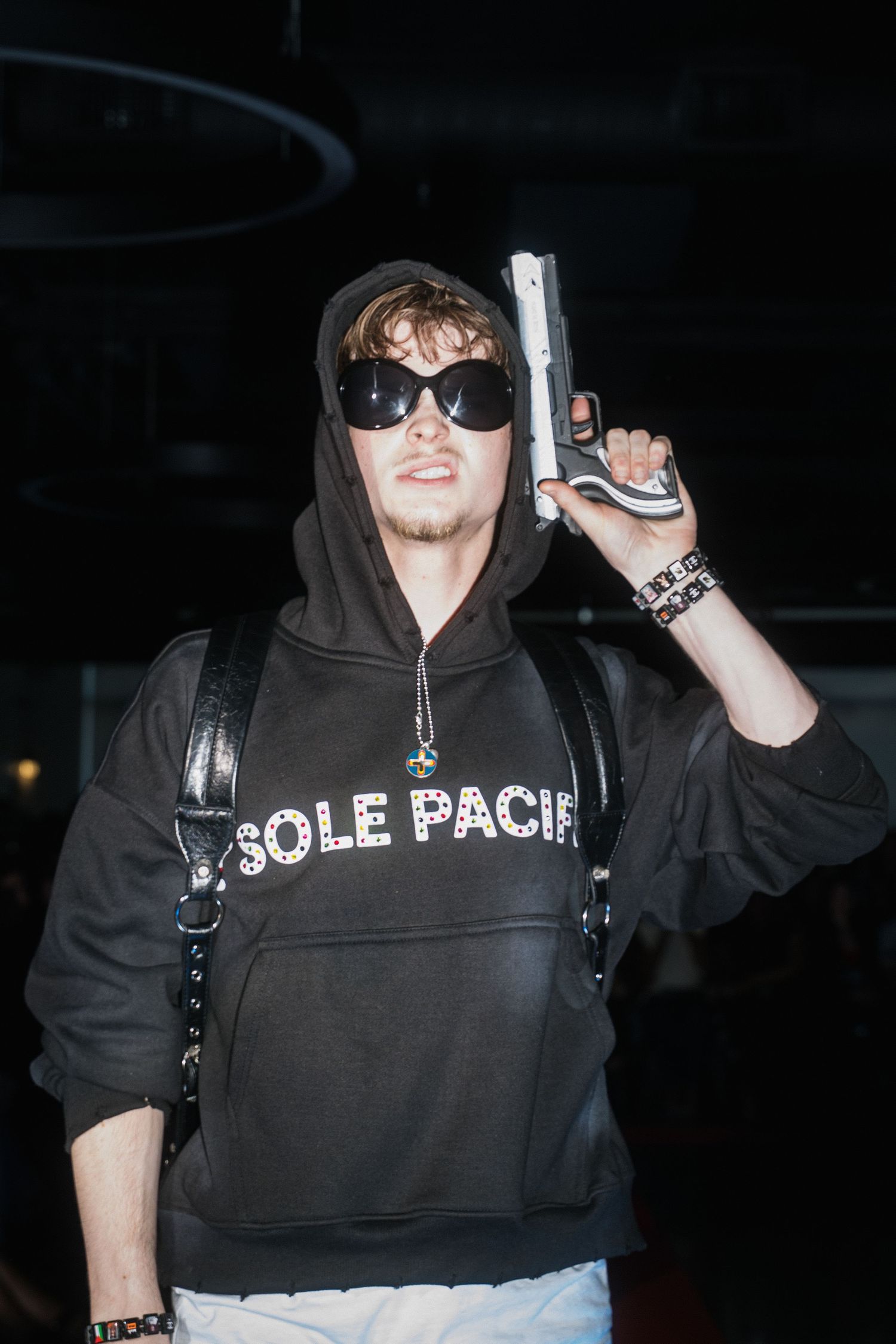 Model in black Sole Pacific hoodie holding prop gun and wearing sunglasses at fashion event.