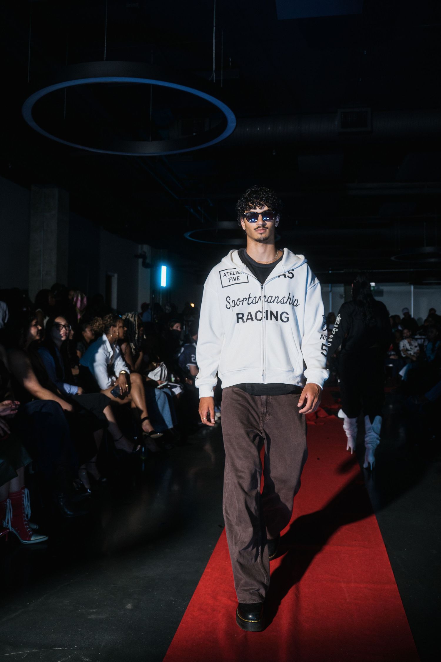 Male model in Sportsmanship Racing white hoodie and brown pants walking a red carpet runway.