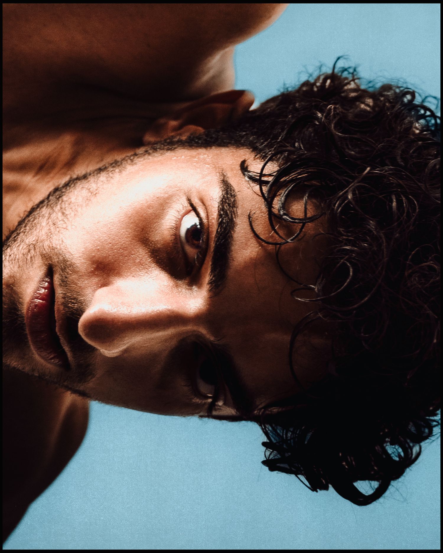 Close-up portrait of a young man with curly dark hair against a blue background.