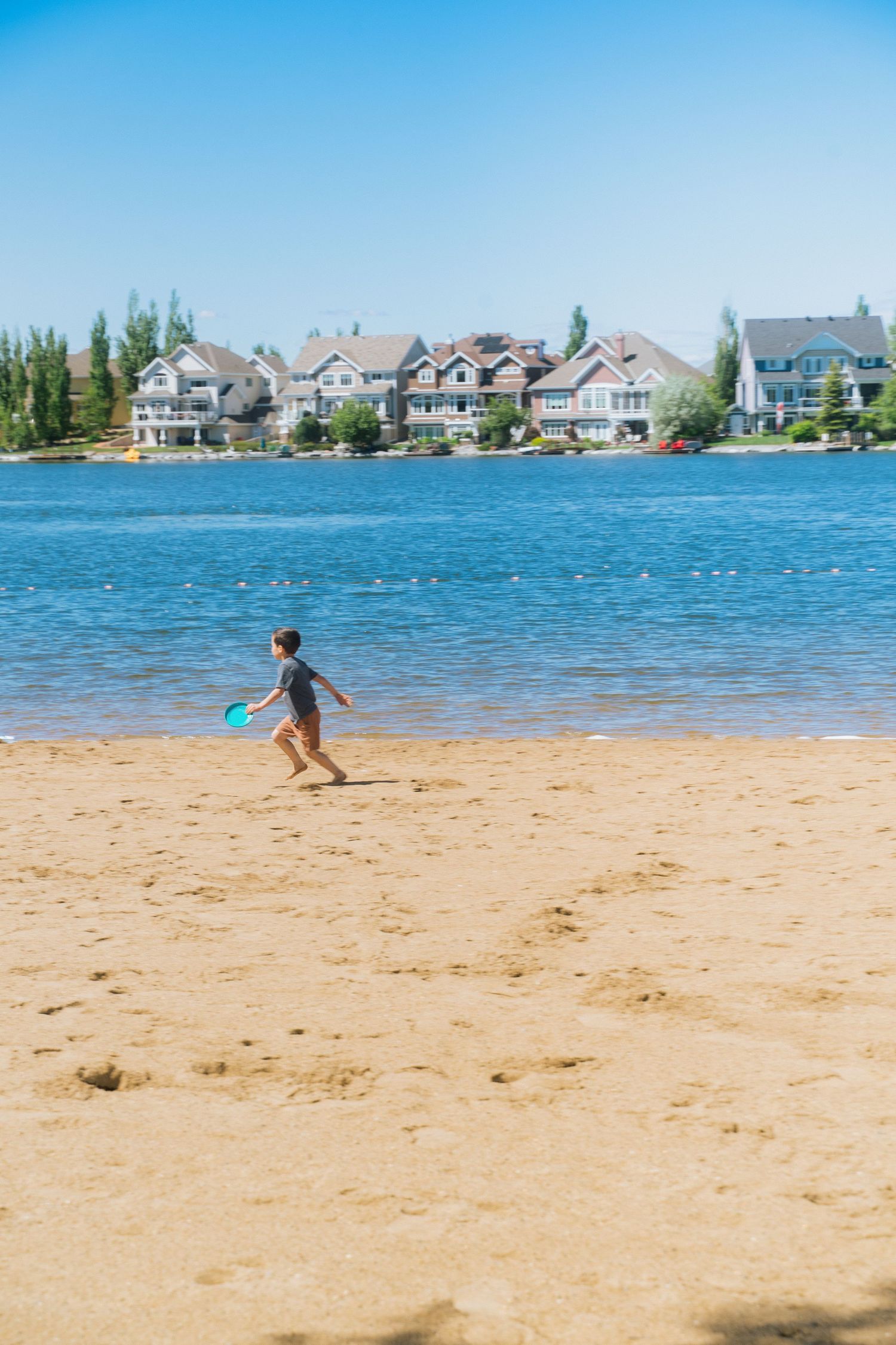 Child playing frisbee on sandy beach with blue lake and luxury waterfront homes in background on sunny day.